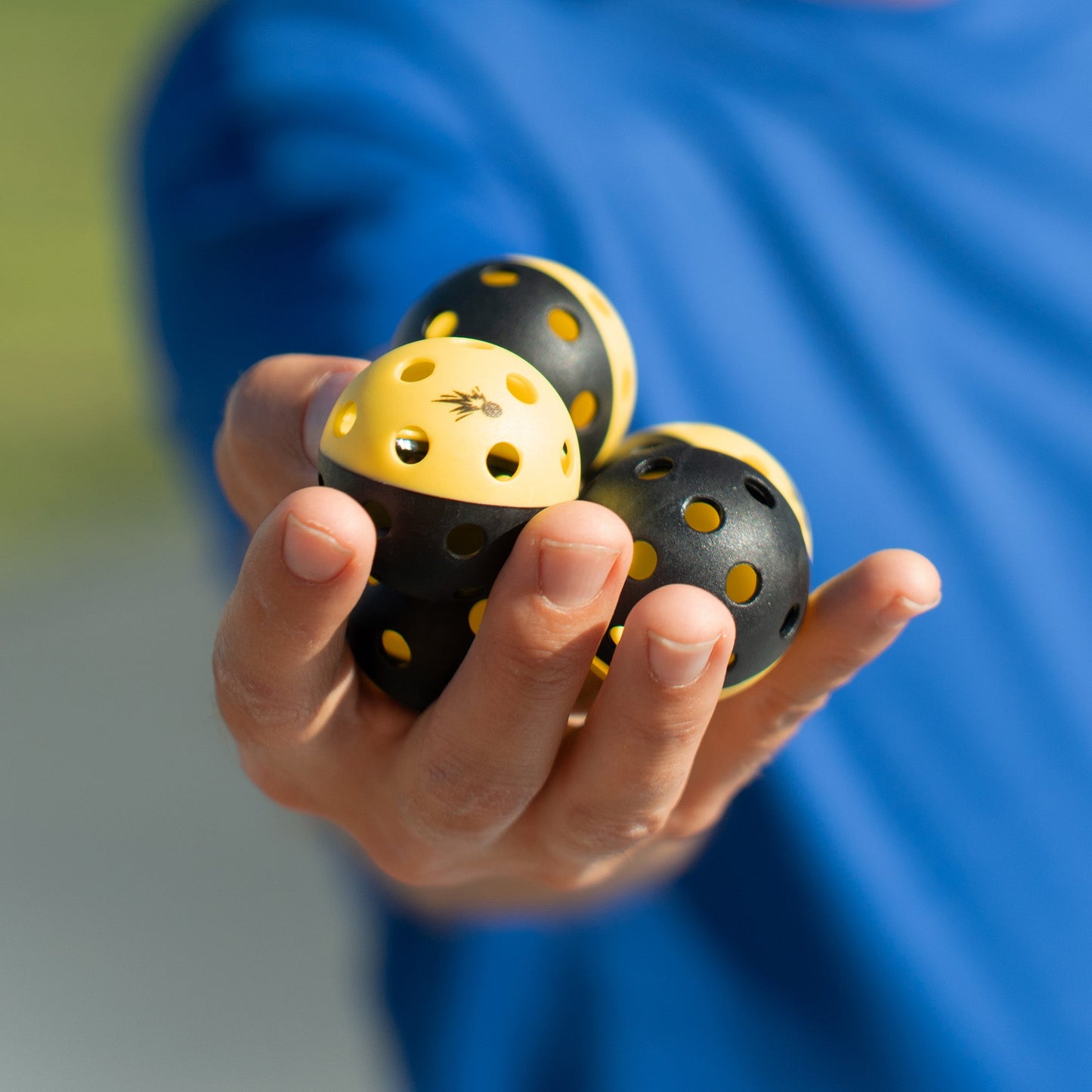 A person in a blue shirt holds out four black and yellow Moment Pickleblasters Mini Plastic Indoor/Outdoor Practice Balls by Moment Pickleball, with the focus on these pickleball training balls designed for skill enhancement.
