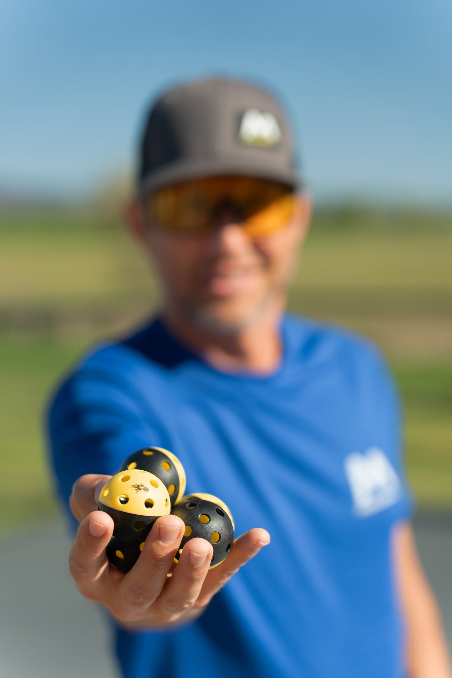 A man in a blue shirt and cap, out of focus, holds four Moment Pickleball’s Pickleblasters 2-Color Mini Plastic Indoor/Outdoor Practice Balls—two yellow, two black—sharply displayed for training against a sunny, blurred background.