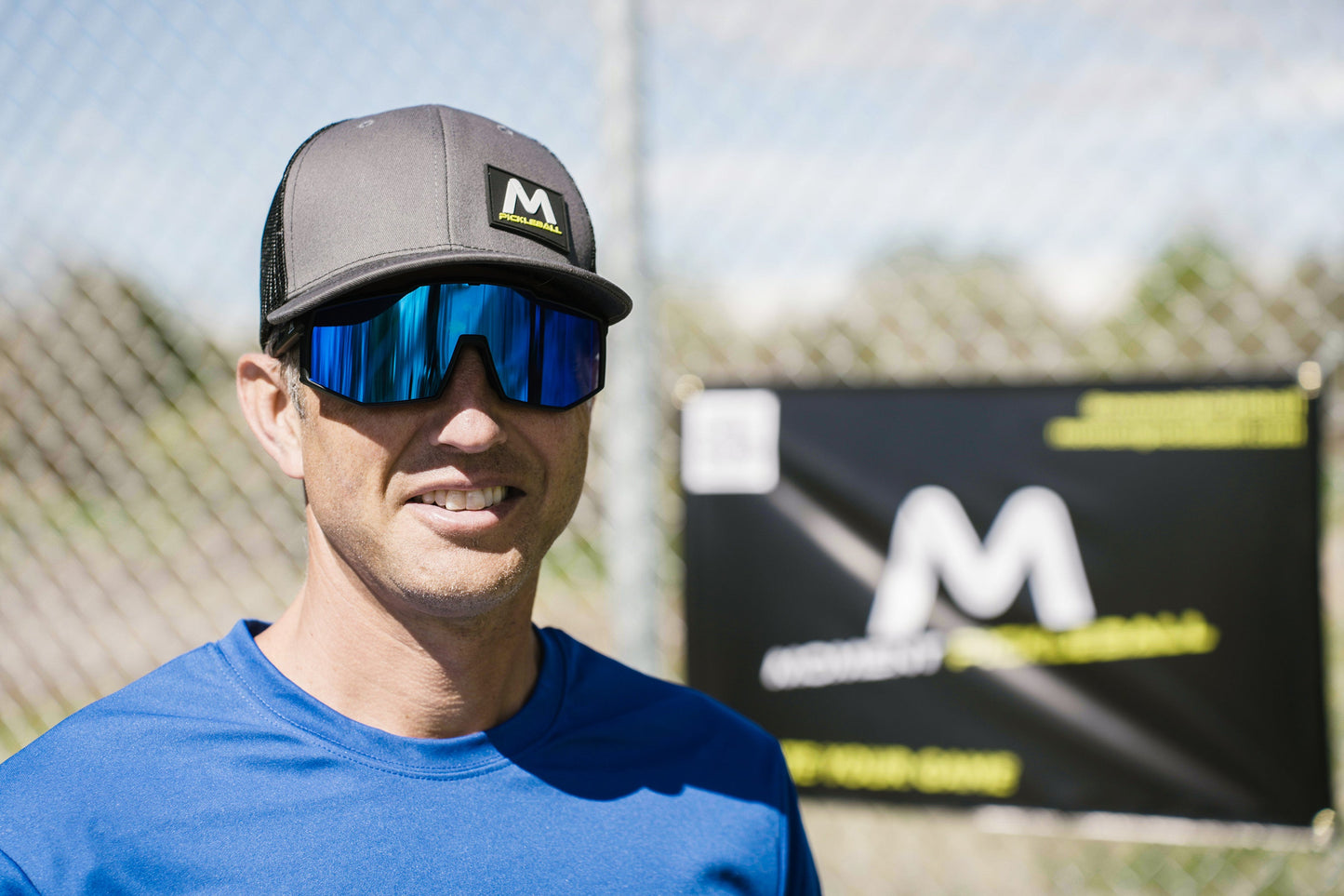 A man wears Moment Pickleball’s Pro-Guard Glasses Set with Blue Mirrored Lens, a blue shirt, and a gray M-logo hat. He stands outdoors in front of a blurred chain-link fence and black sign with a white M.