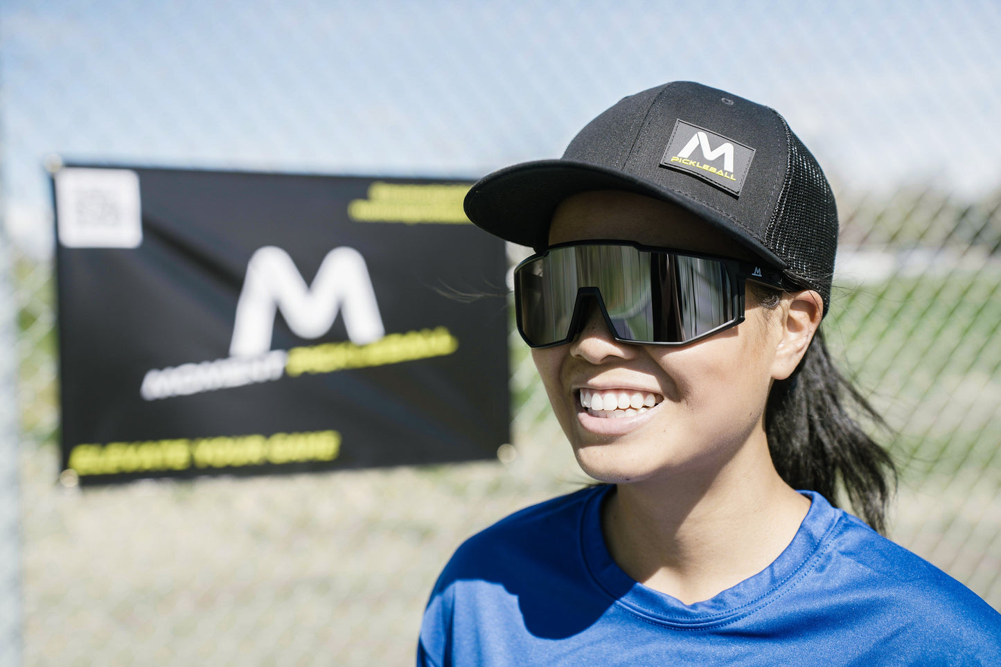Outdoors on a sunny day, a person in a blue shirt and black cap smiles near a chain-link fence displaying a Moment Pickleball banner while wearing the Pro-Guard Glasses Set with Grey Mirrored Lens from Moment Pickleball.