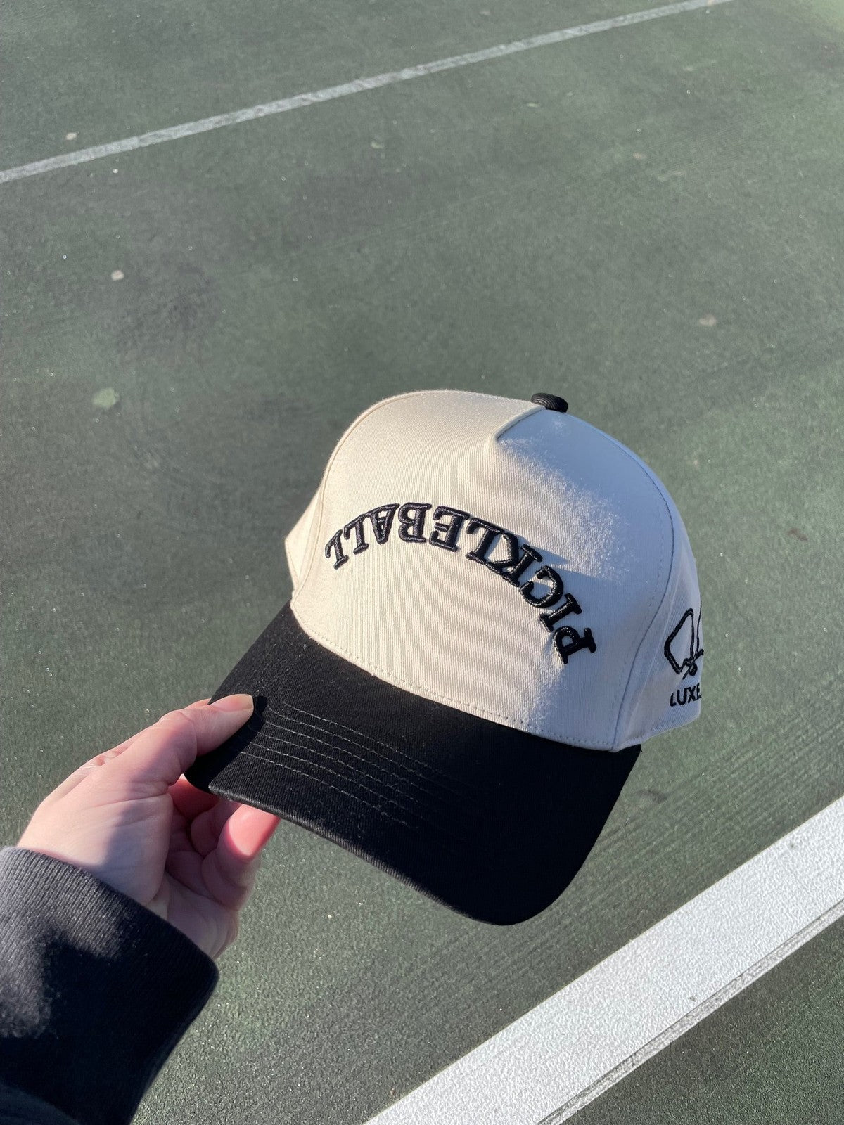 A person holds a beige and black LUXE Pickleball Hat with curved PICKLEBALL text, a small pickleball paddle and ball graphic, LUXE on the side, and sun protection—ideal for outdoor courts.