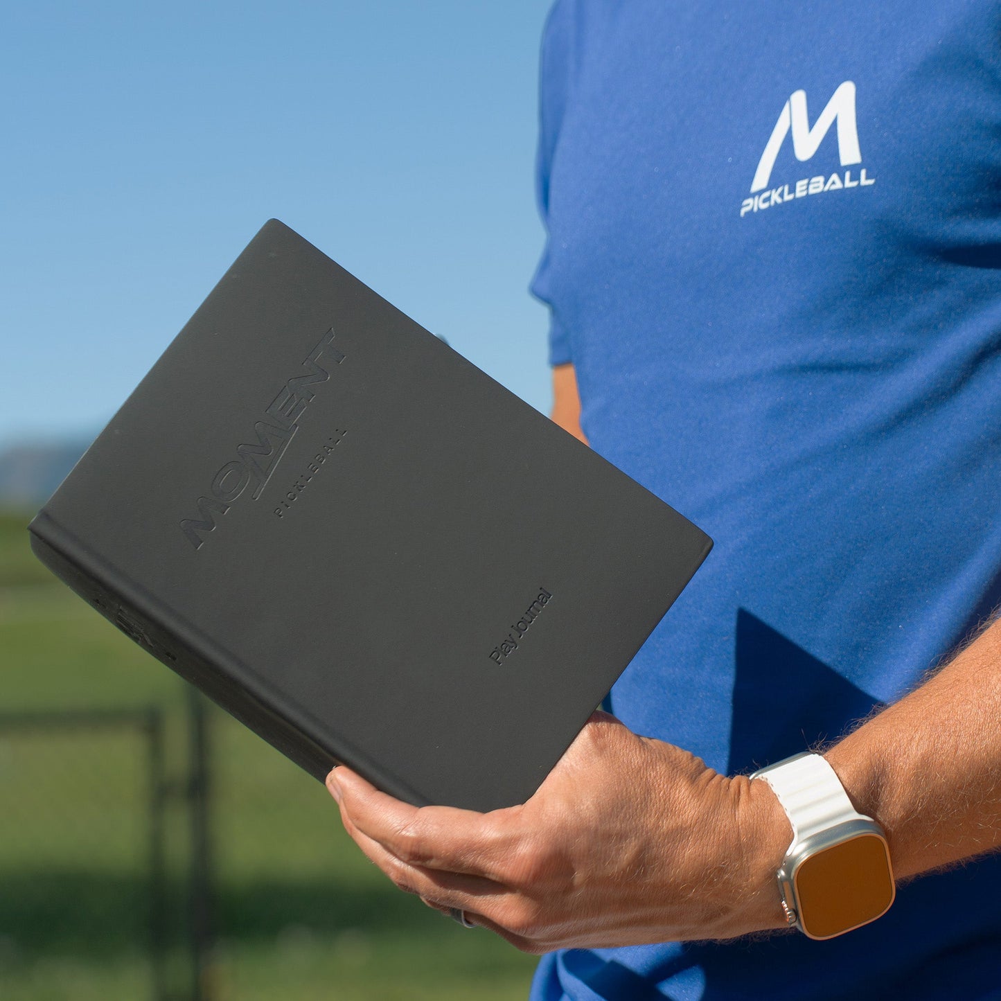 A person in a blue M Pickleball shirt and white smartwatch holds the black Moment Pickleball Practice & Play Mindfulness Journal by Moment Pickleball outdoors, with a blurred fence and clear sky in the background.