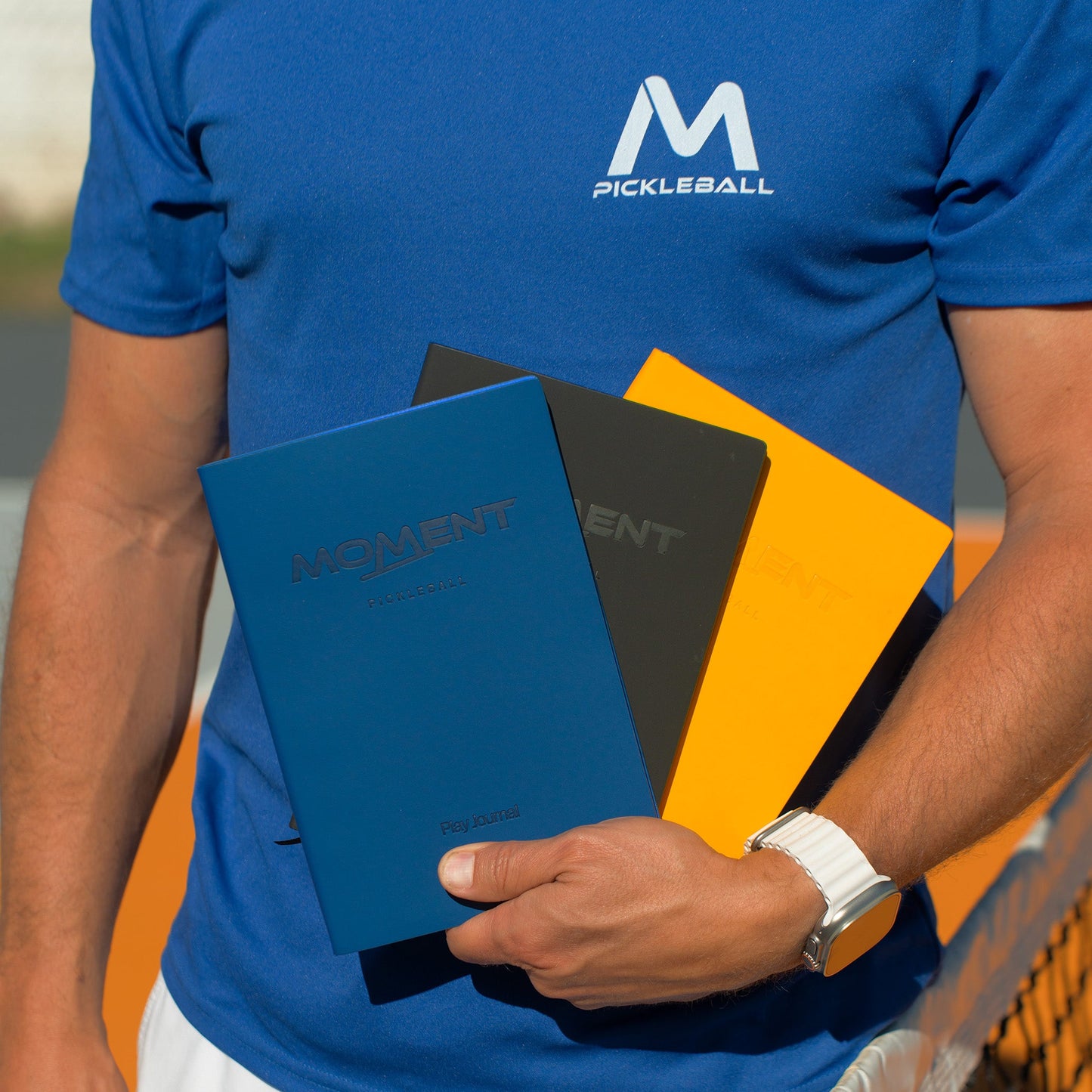 A person in a blue Pickleball shirt holds three Moment Pickleball Practice & Play Mindfulness Journals—blue, black, and yellow editions—by the court net, ready to boost performance with Moment Pickleball’s session-tracking tools.