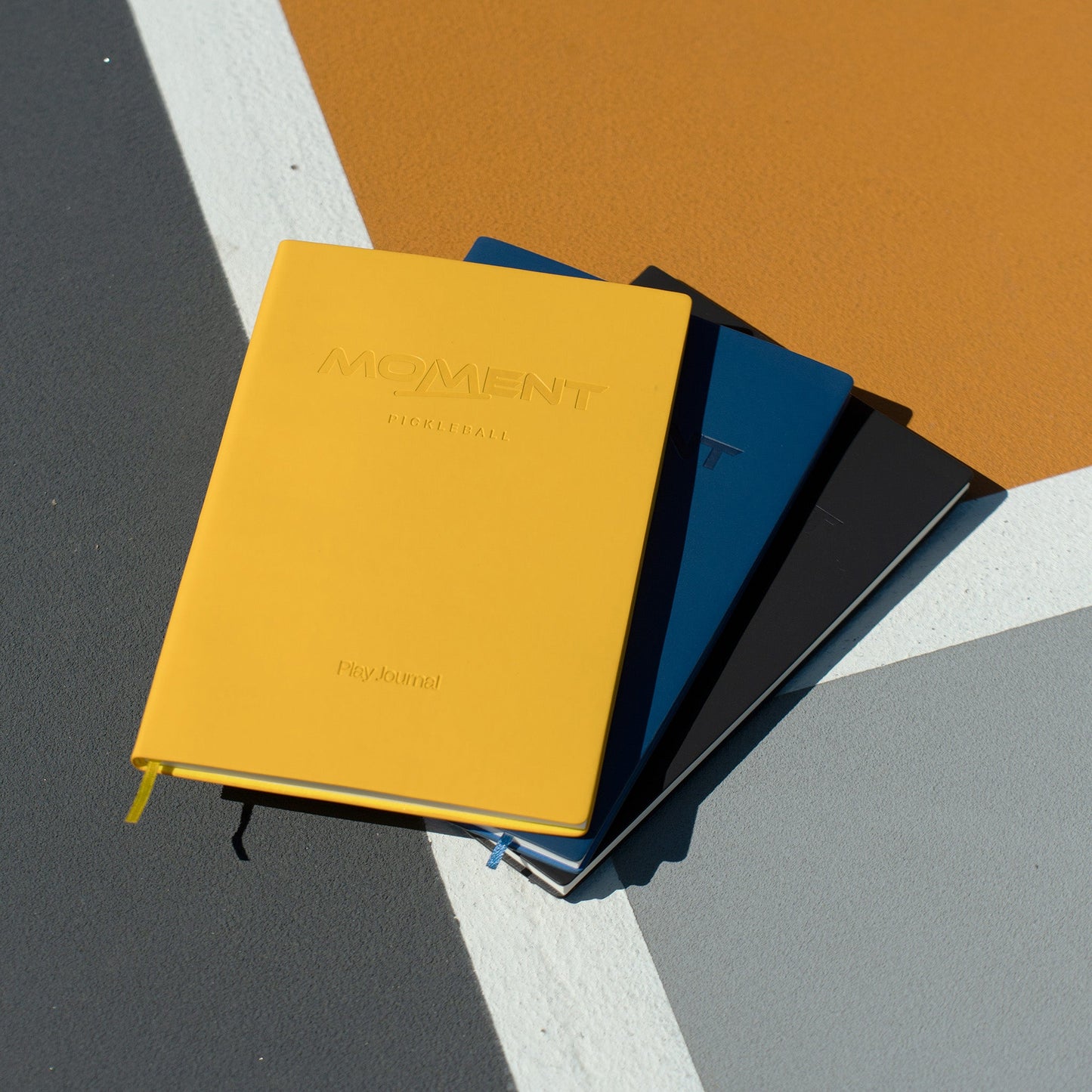 Three hard-cover notebooks—yellow, blue, and black—are stacked on a geometric surface. On top is the Moment Pickleball Practice & Play Mindfulness Journal from Moment Pickleball, ideal for tracking sessions and improving your game.