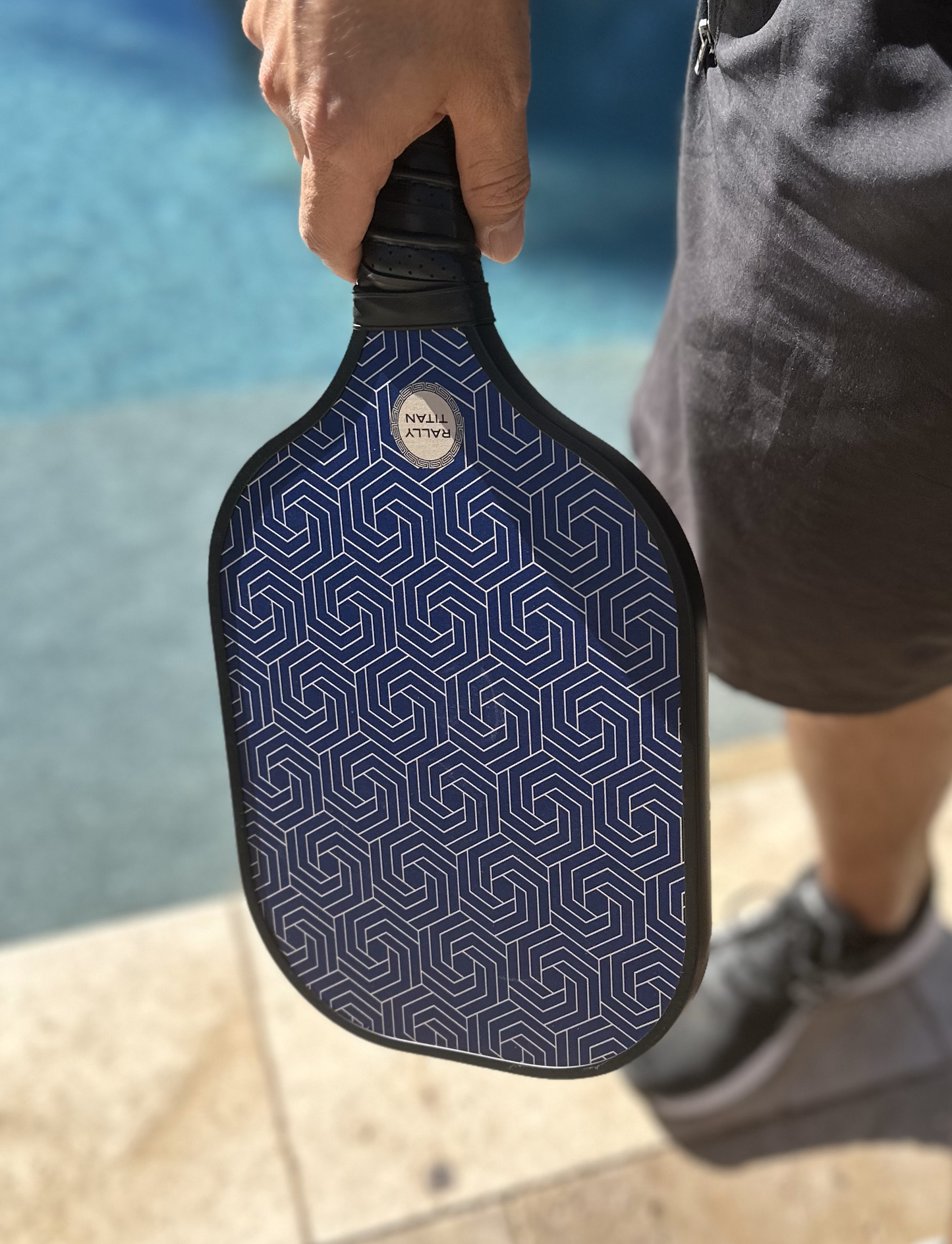 A person stands by a pool holding the Rally Titan Pickleball Blue Hex | 10MM paddle with a T300 textured carbon fiber surface. Sunlight highlights its blue geometric design and the pool’s turquoise water, while they wear dark shorts and gray shoes.