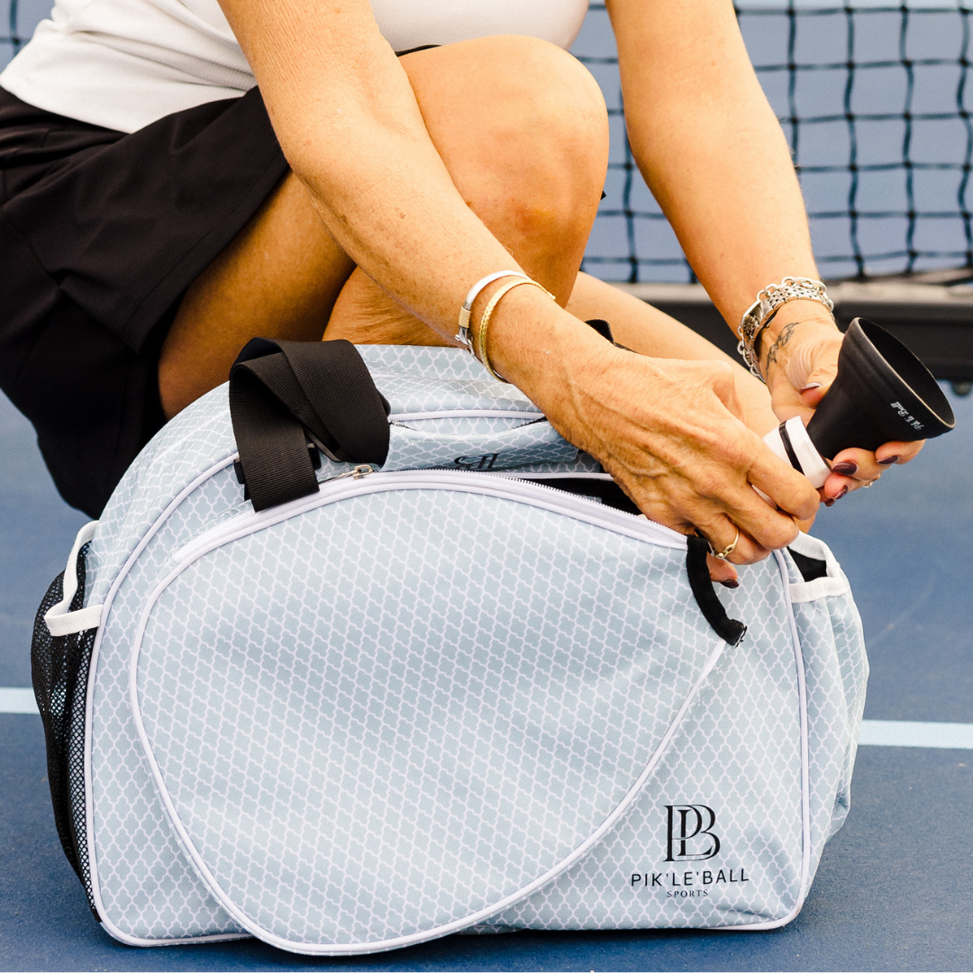Kneeling on an outdoor tennis court, a woman unzips a light gray Pik'le'Ball Sports bag while holding the Pik'le'Ball Ball Retriever, with her pickleball paddle beside her. She wears a black skirt and silver bracelets.
