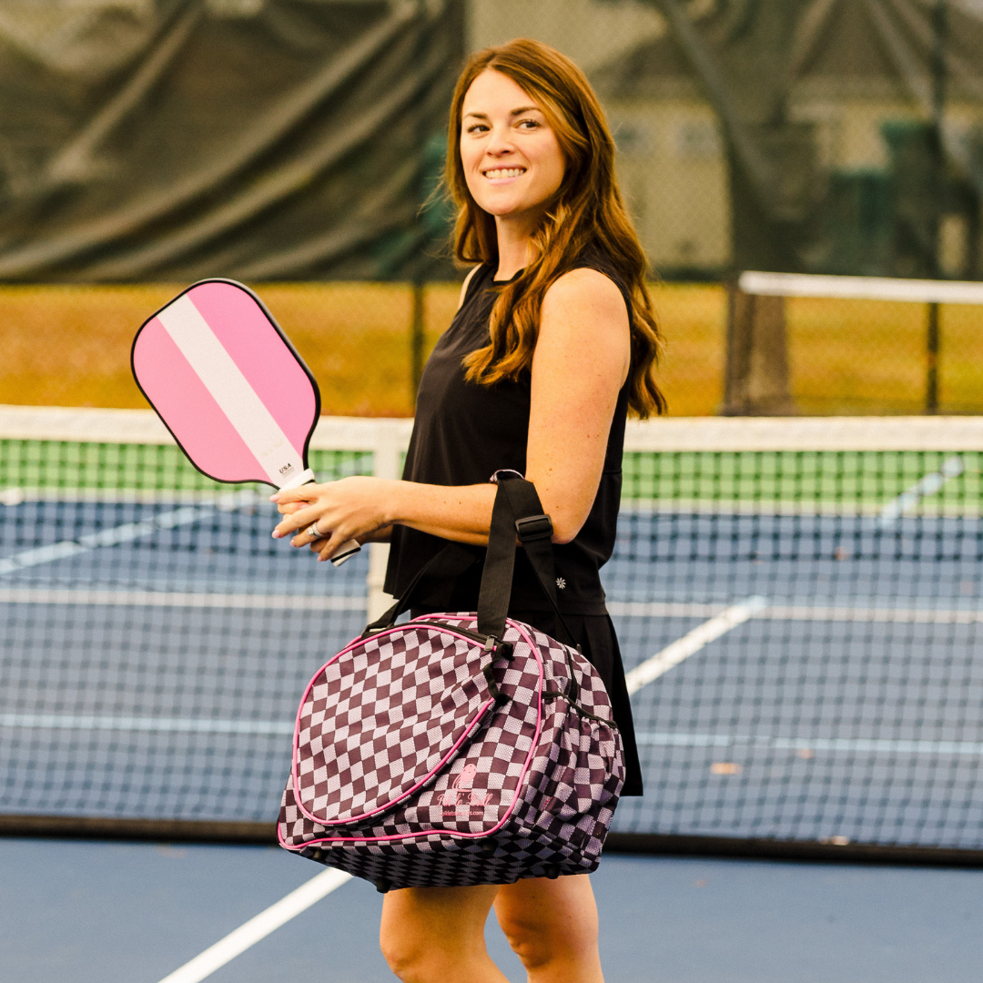 A woman with long brown hair in a black sleeveless outfit stands smiling on a tennis court, holding a pink and white paddle. She carries the Checkered Chic Black Premium Women's Pickleball Bag by Pik'le'Ball Sports over her shoulder.