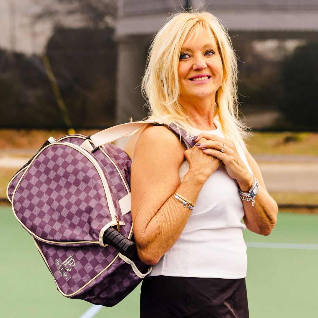A blonde woman in a white sleeveless top and dark skirt stands on a tennis court, smiling as she carries the Pik'le'Ball Sports Checkered Chic Premium Women's Pickleball Bag with padded compartments and adjustable strap over her shoulder.