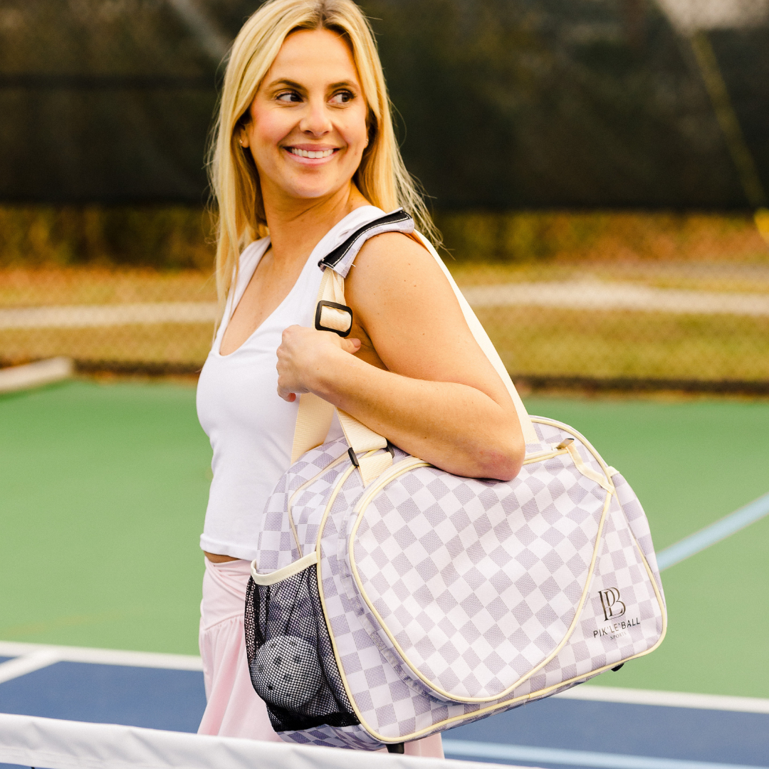 A smiling blonde woman carries the Pik'le'Ball Sports Checkered Chic Premium Women's Pickleball Bag with padded compartments and adjustable strap over her shoulder on a court, wearing a white tank top and pale pink pants.
