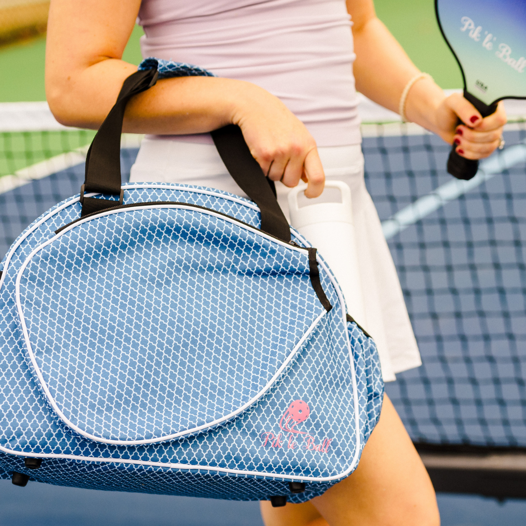 A woman in a light purple top and white skirt holds the Pik'le'Ball Sports Classy Premium Women's Pickleball Bag, a stylish blue organizer with a white geometric pattern, black straps, and space for gear including her paddle and water bottle.