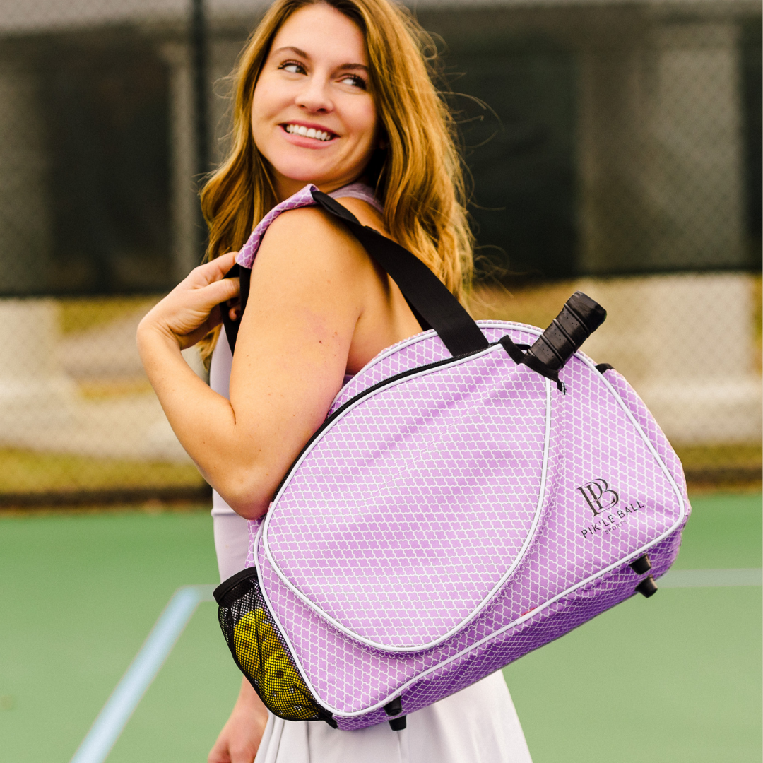 A smiling woman with long light brown hair stands on a tennis court, holding the Pik'le'Ball Sports Classy Premium Women's Pickleball Bag—a stylish organizer with paddle and gear storage—over her shoulder. She wears a sleeveless white dress by the net.