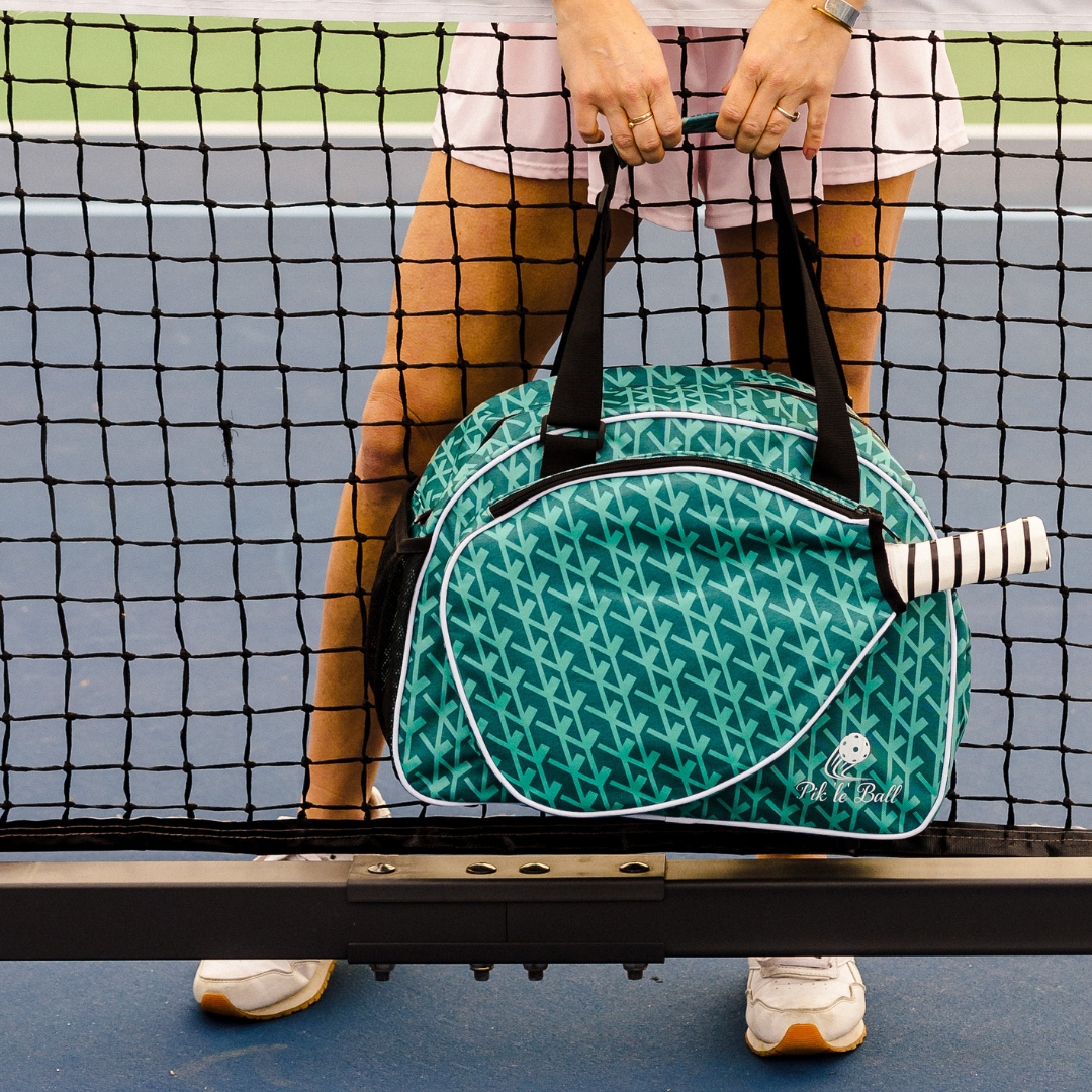 A person in tennis attire stands behind a net, holding the Pik'le'Ball Sports Court Couture Premium Women's Pickleball Bag with multiple compartments and a paddle sleeve. The blue court highlights their white skirt and sneakers.