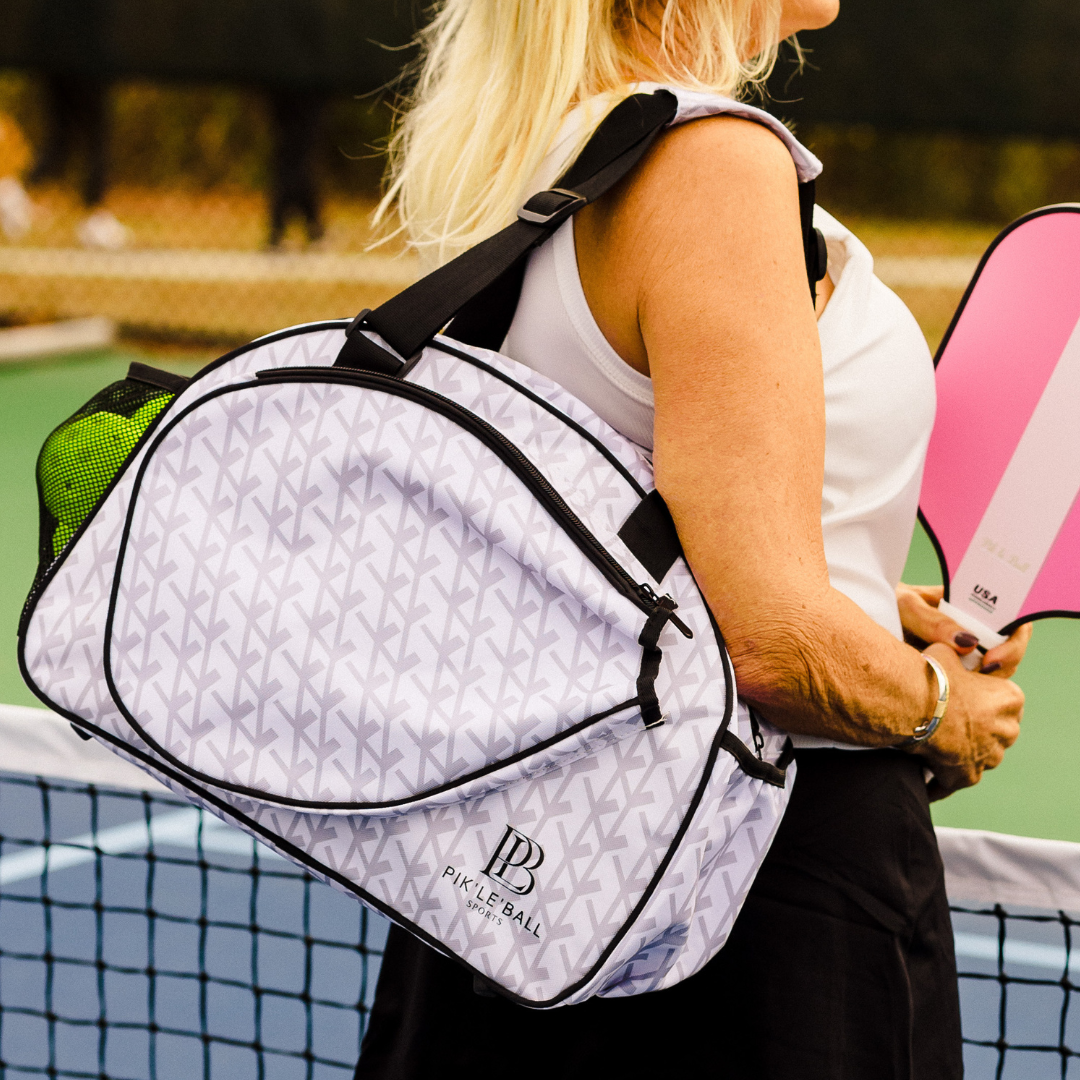 A woman in a white sleeveless top carries the Pik'le'Ball Sports Court Couture Premium Women's Pickleball Bag with multiple compartments and paddle sleeve on her shoulder, holding a pink paddle by an outdoor tennis net amid blurred trees and fencing.