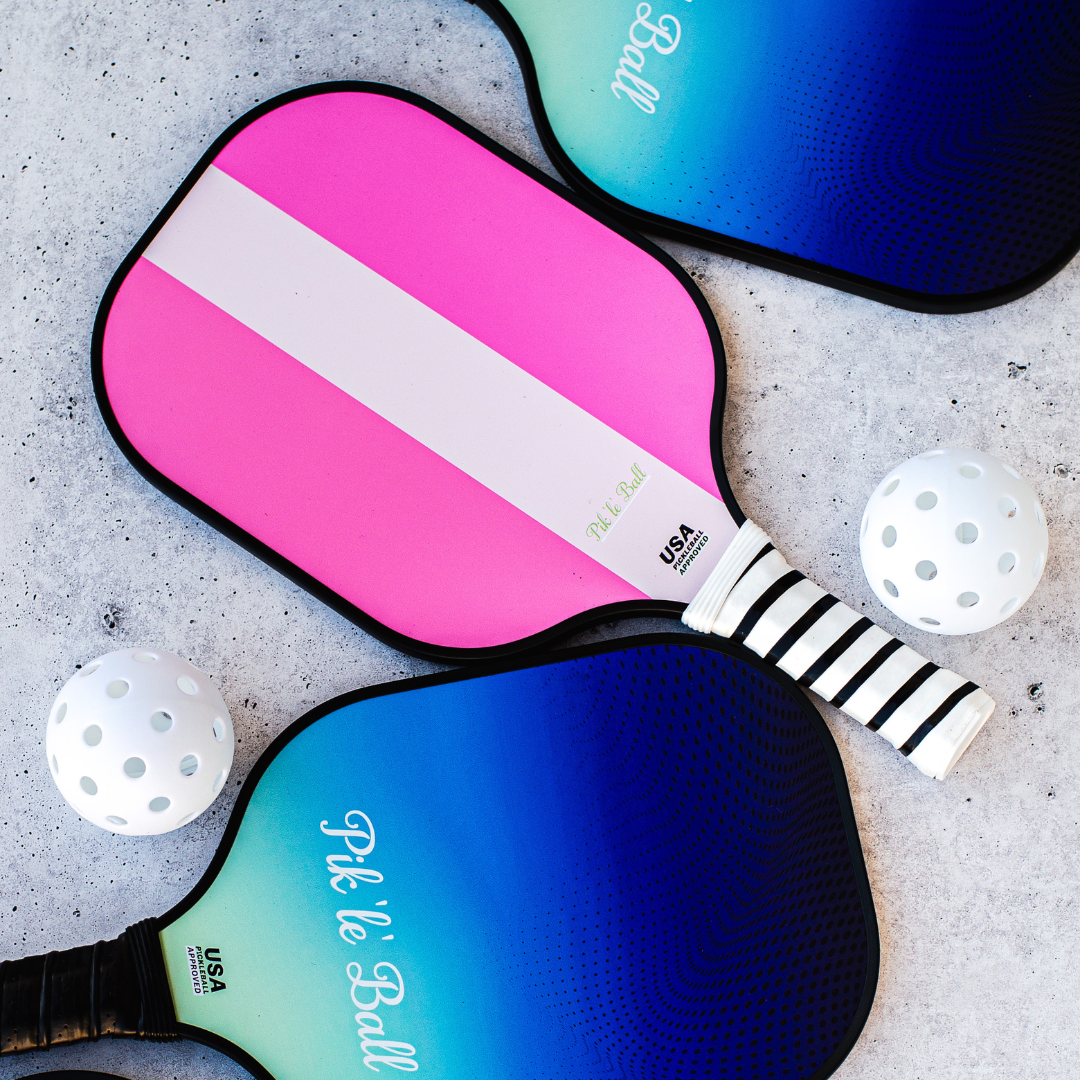 Two USAPA approved Carbon Fiber Pickleball Paddles from Pik'le'Ball Sports—one pink with a white stripe, one blue gradient—rest on a light textured surface, branding and USA Pickleball logos visible, with three white pickleballs nearby.