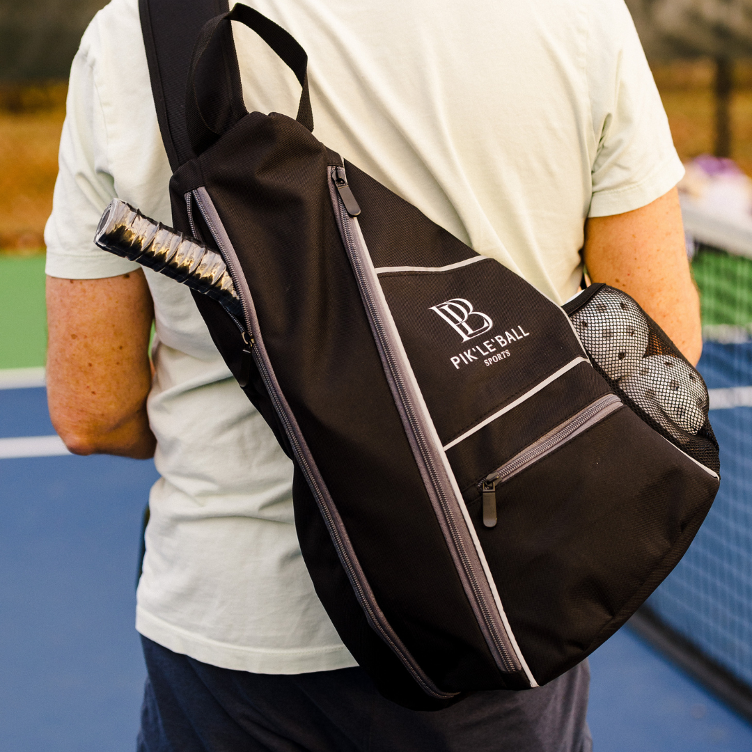 A person in a light green shirt stands on a blue and green court, carrying the Pik'le'Ball Sports Sporty Sling Grey Pickleball Bag, with a paddle handle sticking out and a mesh pocket for a water bottle. The net and autumn leaves are in the background.