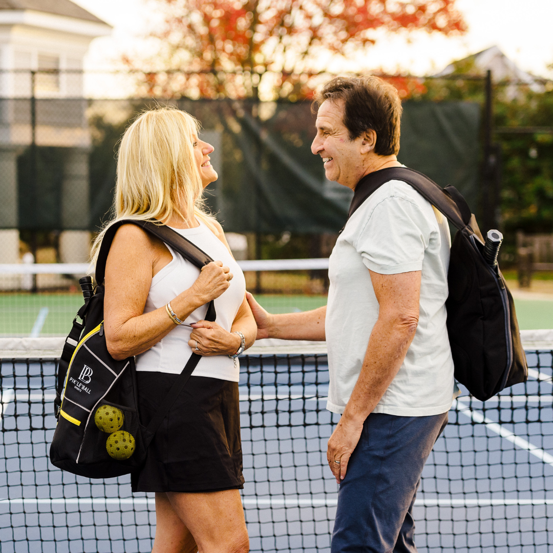 A smiling older couple stands on a tennis court holding Pik'le'Ball Sports Sporty Sling Yellow Pickleball Bags and paddles over their shoulders, separated by the net, with autumn trees and orange leaves in the background.