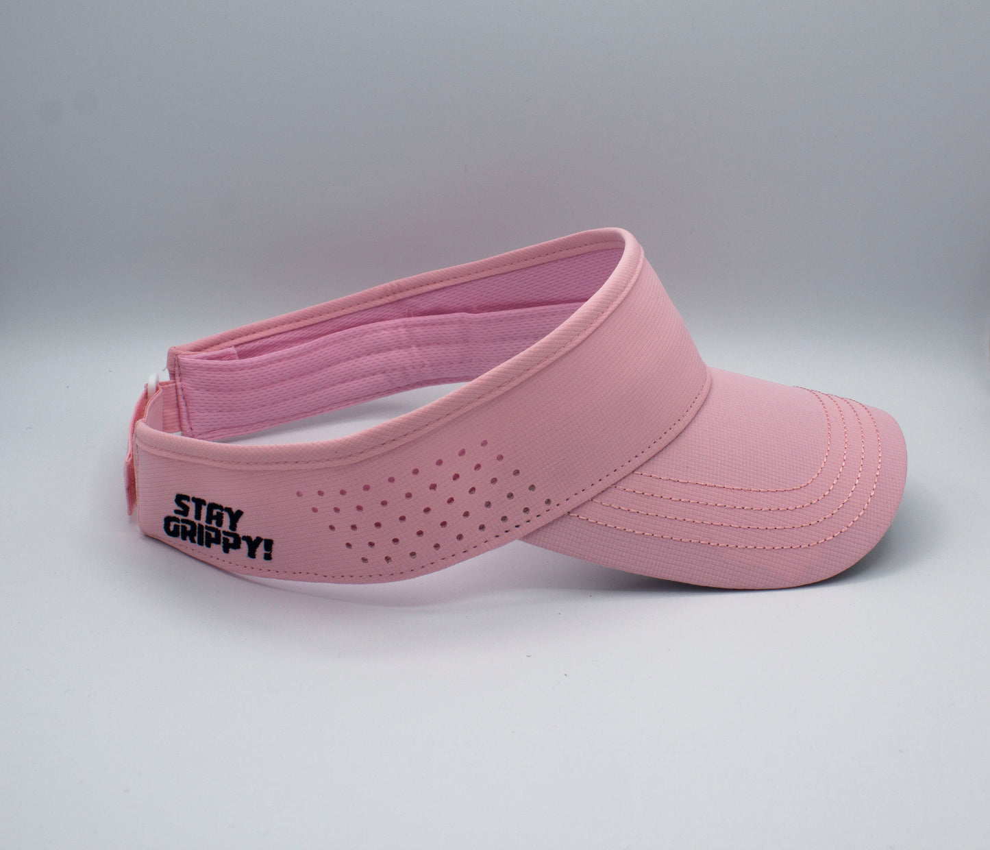 The Bodhi Performance ICON Visor is a light pink performance visor with small side ventilation holes, “STAY GRIPPY!” in black on the left, visible brim stitching, and an adjustable back strap, displayed against a white background.