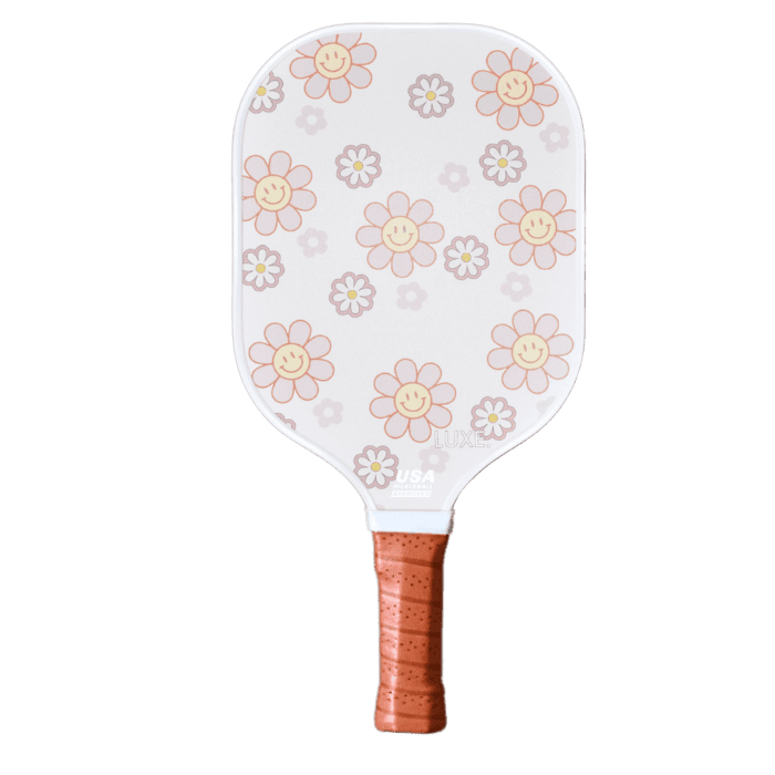 The LUXE Pickleball Smiley Flower paddle is USA Pickleball Approved and ideal for beginners, featuring cartoon pink and orange smiling flowers with yellow centers and white daisies on a light gray background, plus a brown leather grip.