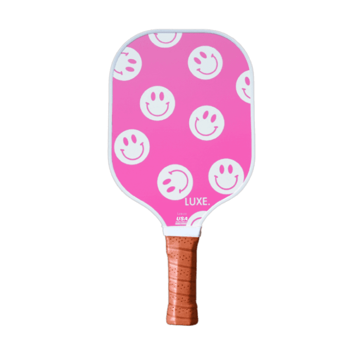 The LUXE Pickleball Smiley paddle features a pink fiberglass surface with white smiley faces, a brown perforated grip, “LUXE.” in white at the bottom edge, and is USA Pickleball Approved. The background is plain white.