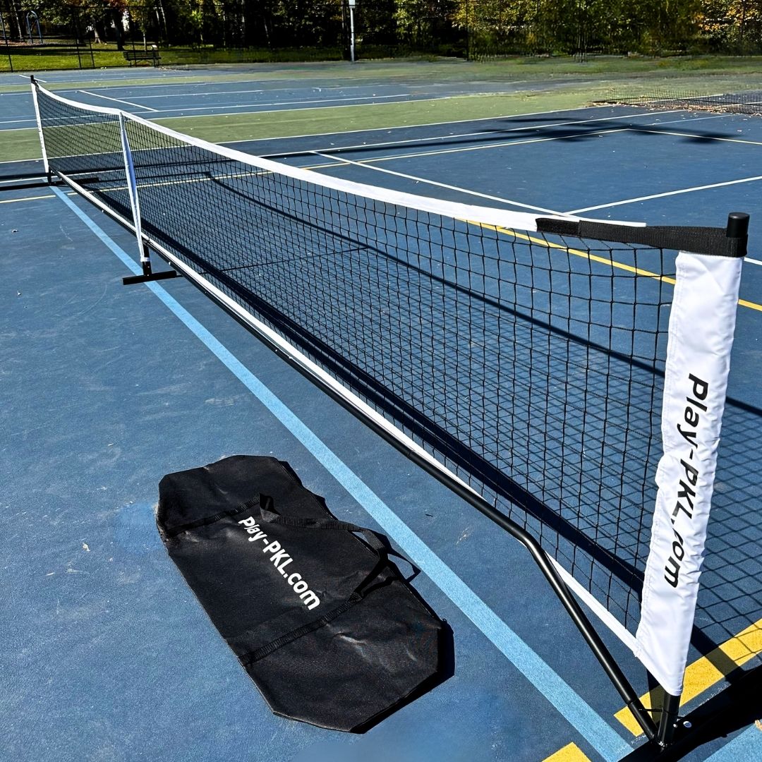 The Playly Store Logo Portable Pickleball Net is set up on a blue outdoor court. The net posts and black carrying bag feature play-pkl.com branding. Trees and empty courts can be seen in the background.