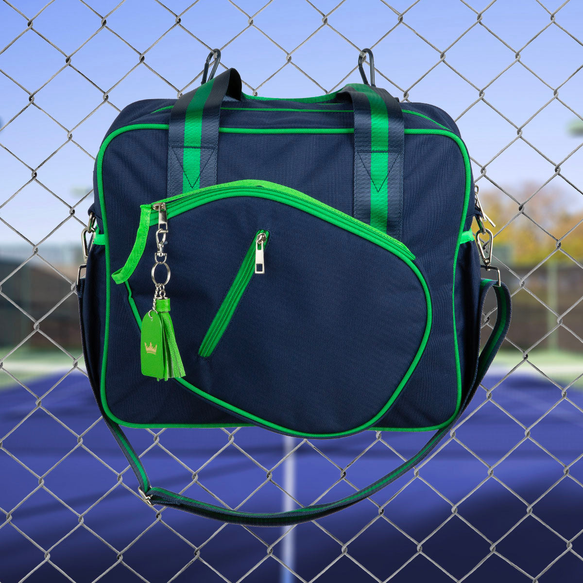 The Queen of the Court Prepster Pickleball Bag in navy blue with green zippers and trim hangs on a chain-link fence by a tennis court. It includes two handles, a shoulder strap, and a keychain with a green tassel and silver crown charm.