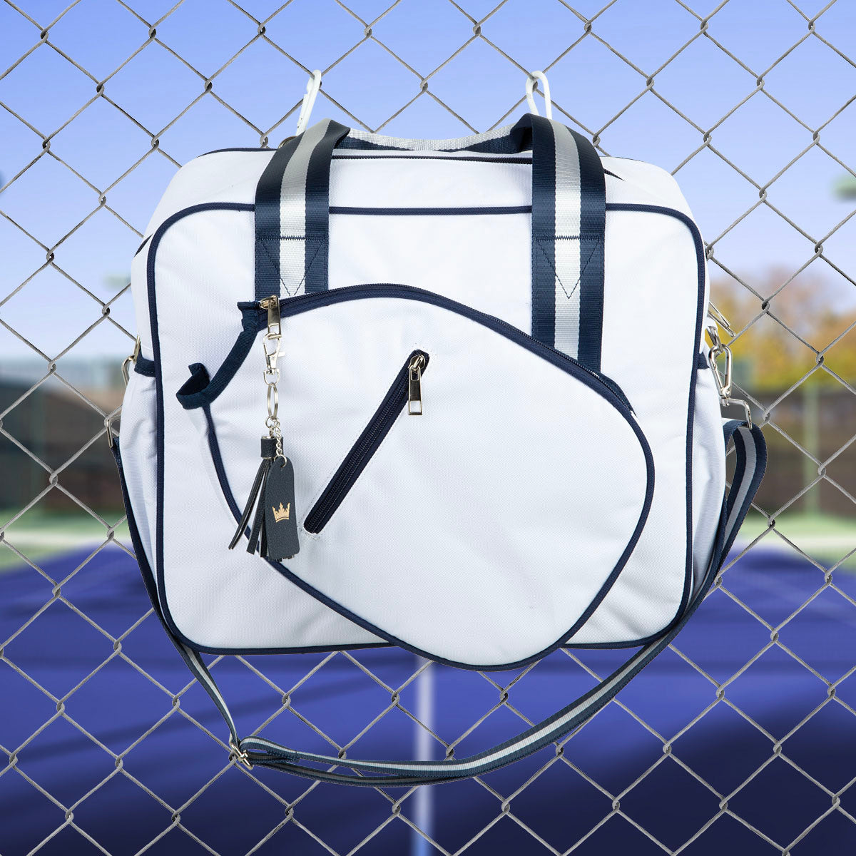 The Queen of the Court Prepster Pickleball Bag in white with navy trim and handles hangs on a chain-link fence, showcasing its zippered front pocket and gold crown charm. A blue tennis court and blurred trees are visible in the background.
