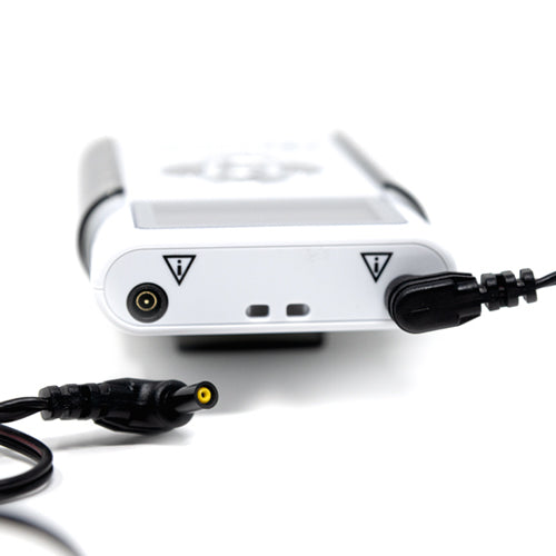 Close-up of the iReliev TENS + EMS Pain Relief & Recovery System, showing two ports (circular and rectangular) with warning triangle symbols, plus black cables—one connected, one loose—against a plain white background.