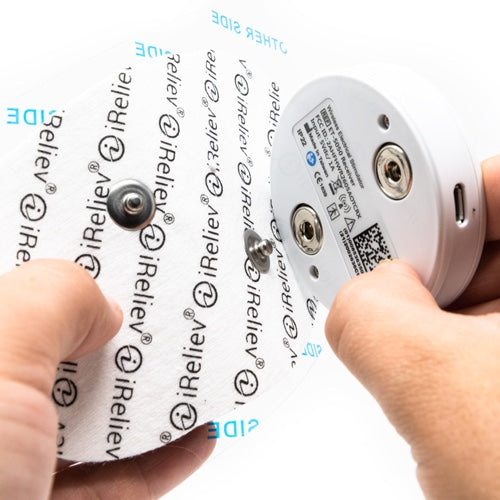 Two hands attach a white, circular iReliev electrode pad to the Therapeutic Wearable System. The pad connects via TENS technology to the device, which features a QR code and alignment instructions for targeted pain relief.