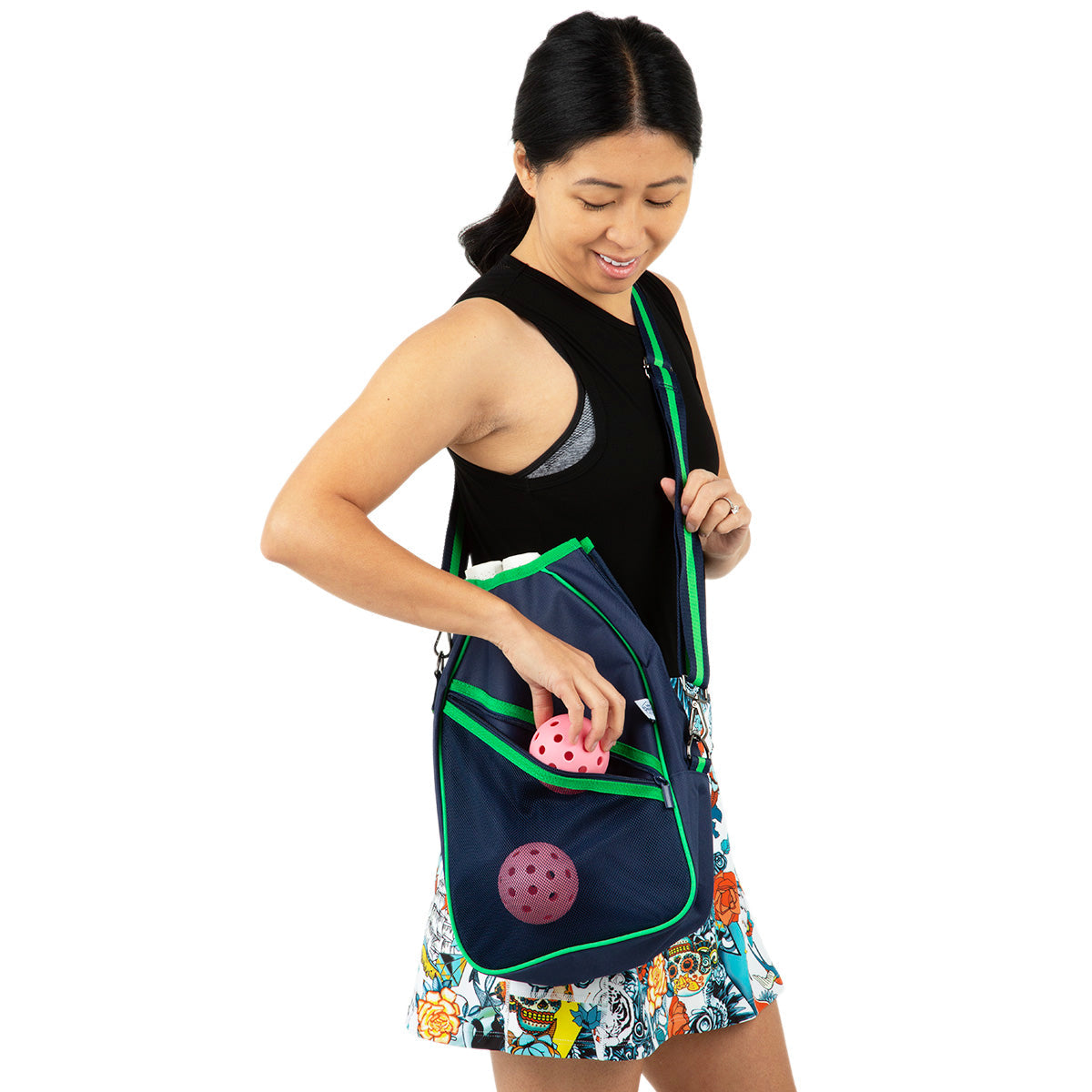 A woman in a black sleeveless top and floral skirt smiles as she places a pink pickleball into her blue and green Queen of the Court Essentials Pickleball Bag, which has another pink ball in the front mesh pocket.