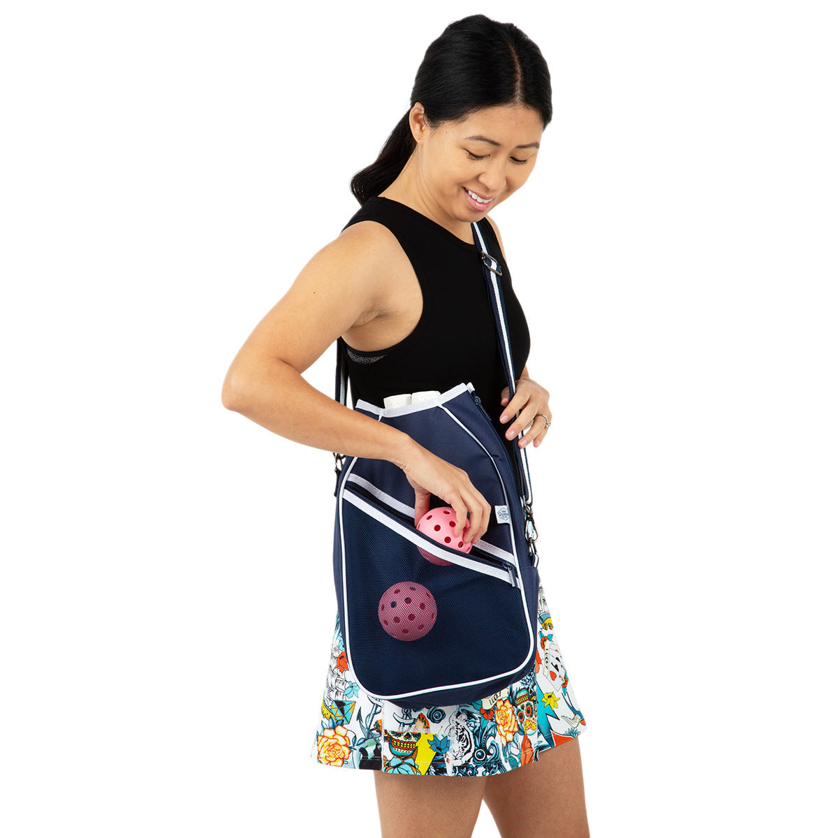 A woman in a black top and floral skirt smiles as she puts a pink perforated ball into her Queen of the Court Essentials Pickleball Bag, a navy crossbody with white trim and an outer mesh pocket holding more pickleball gear.