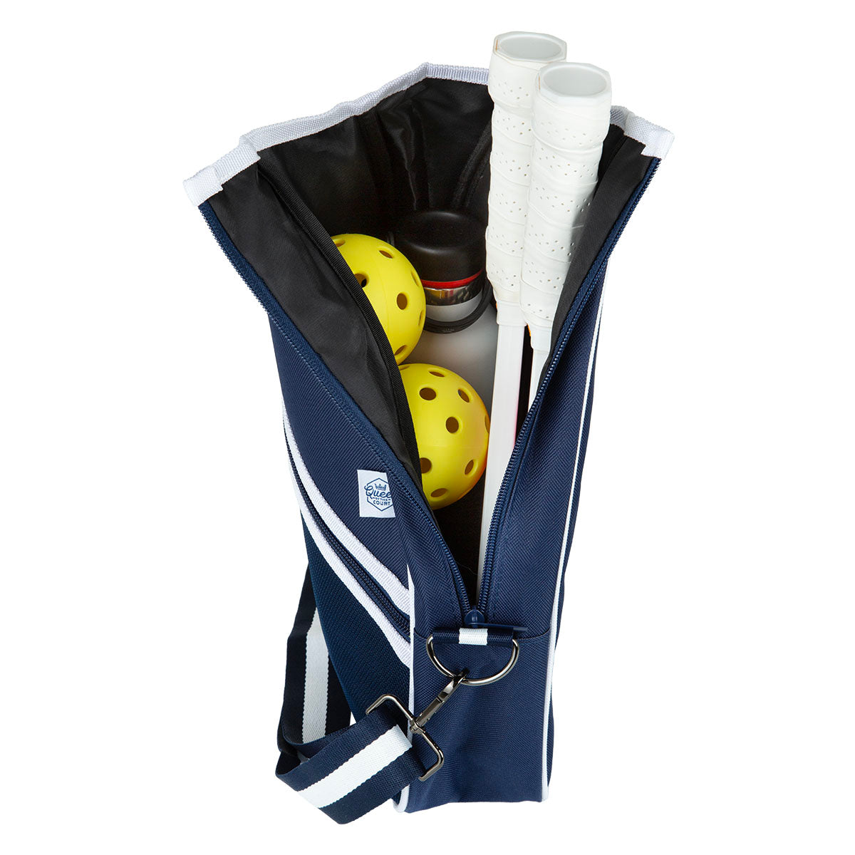 The Queen of the Court Essentials Pickleball Bag is a navy blue crossbody with white stripes, a front logo patch, and shoulder strap. Unzipped, it displays pickleball gear: two yellow balls, two white-handled paddles, and a small black container.