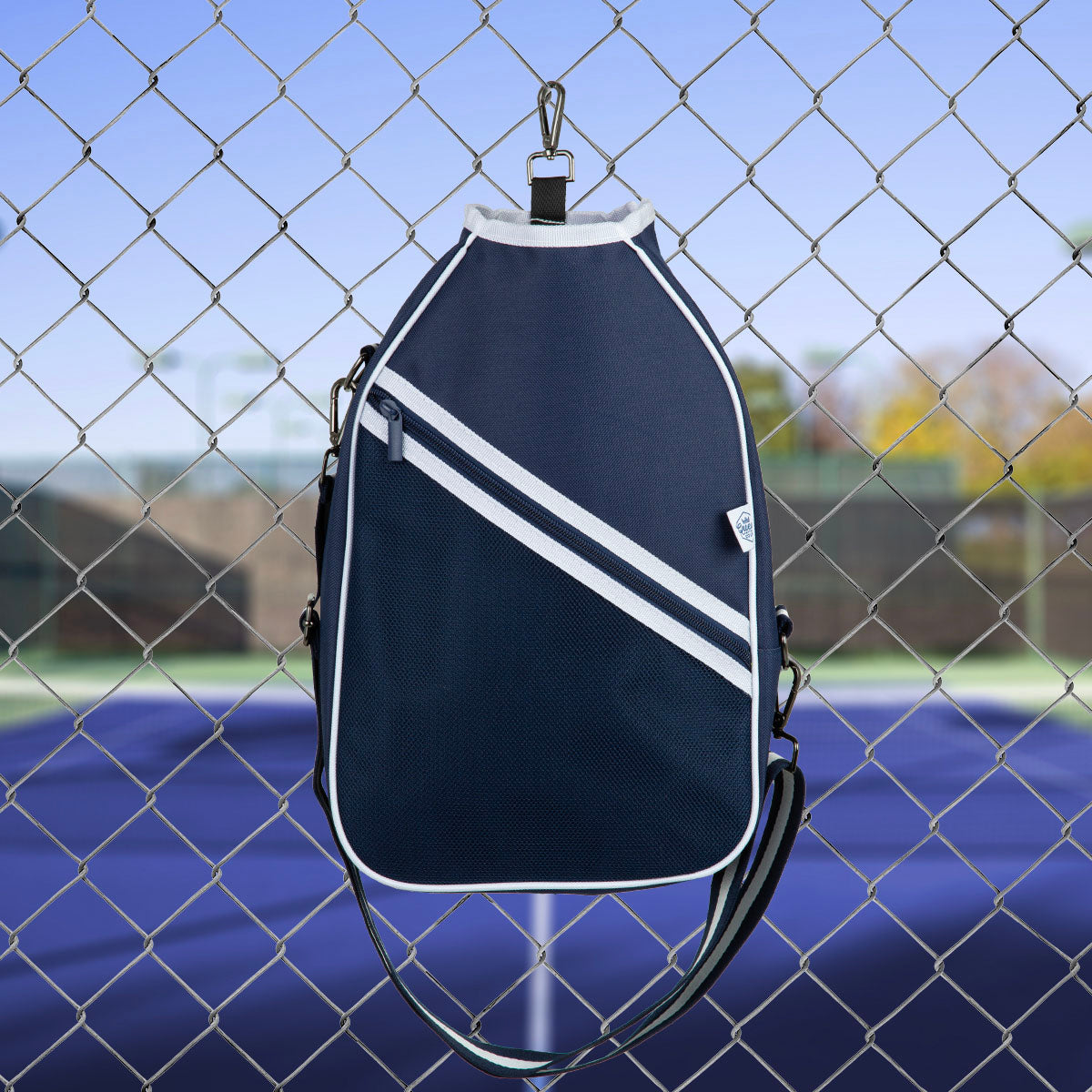 The Queen of the Court Essentials Pickleball Bag in navy blue with white trim and diagonal stripes hangs on a chain-link fence, with a blue tennis court and trees softly blurred in the background to showcase this stylish accessory.