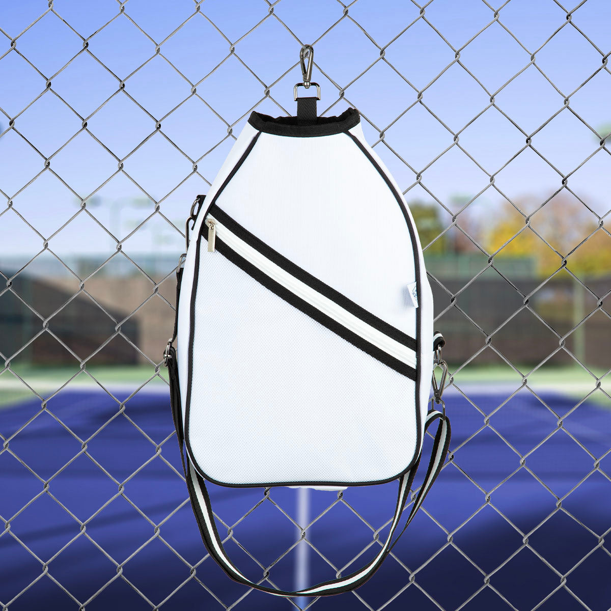 The Queen of the Court Essentials Pickleball Bag, a white crossbody with black accents and a diagonal stripe, hangs on a fence by a blue court. It features a shoulder strap and small zipper pocket—ideal for your pickleball essentials.