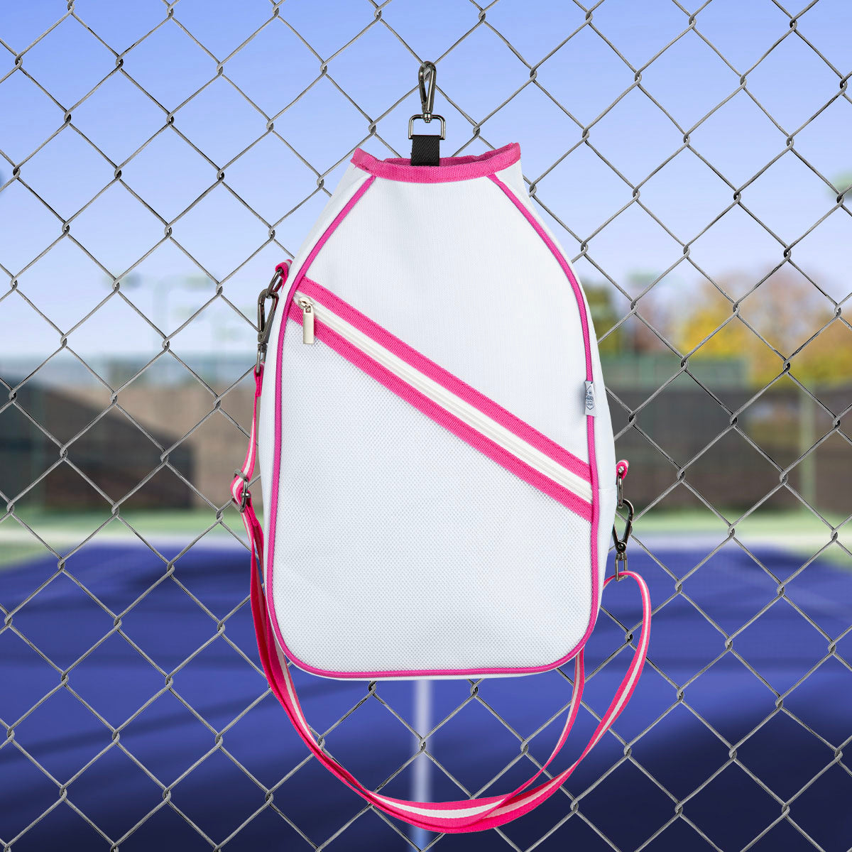 The Queen of the Court Essentials Pickleball Bag, in white with pink accents and a pink strap, hangs on a chain-link fence above a blue tennis court. This crossbody bag features silver hardware and a diagonal zippered pocket.