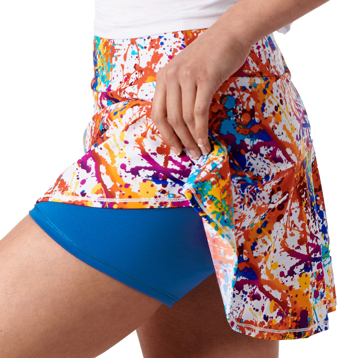 A person lifts the side of a Queen of the Court Splatter Paint Swing Skirt, revealing blue shorts underneath. The colorful skirt features a splatter paint pattern and is paired with a white top; only their lower torso and right hand are visible.