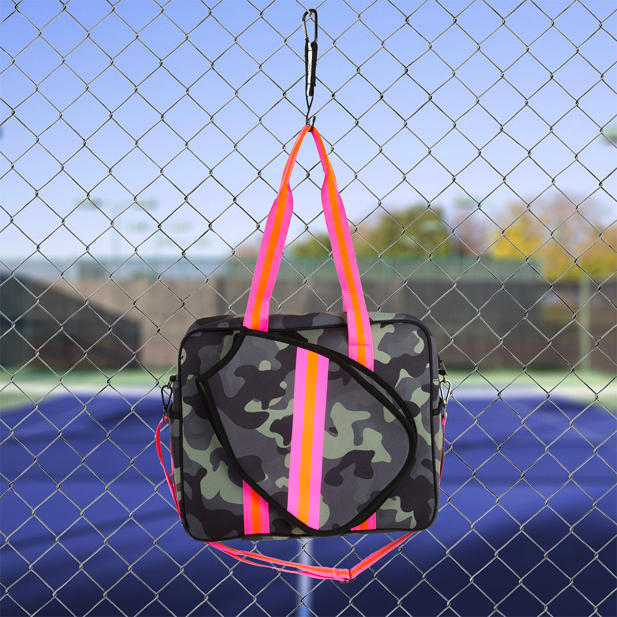 The Queen of the Court Brooklyn Pickleball Bag, featuring a camouflage neoprene design with bold pink and orange straps, hangs on a chain-link fence beside a blue tennis court under a clear sky.
