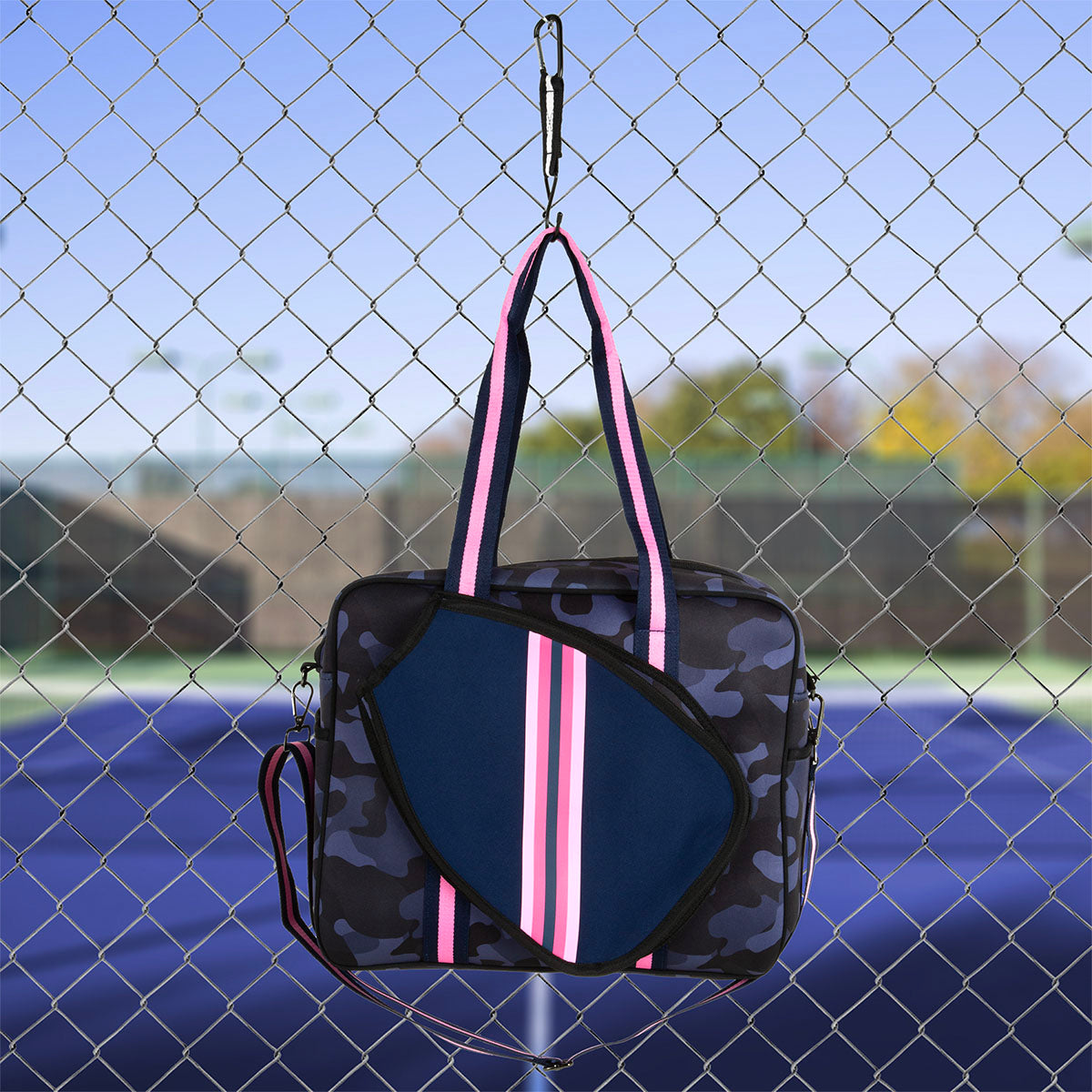 The Queen of the Court Brooklyn Pickleball Bag, featuring a blue camouflage pattern and pink, white, and navy striped handles, hangs on a chain-link fence with a blurred blue tennis court and greenery in the background.