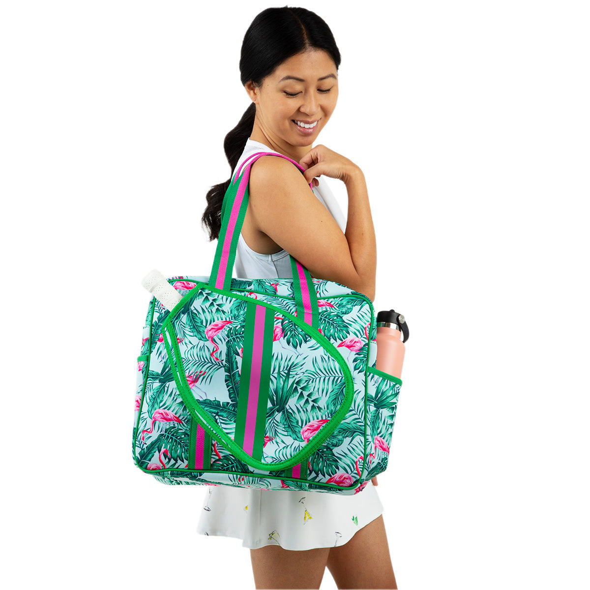 A woman with long dark hair in a ponytail and a sleeveless white dress smiles as she carries the Queen of the Court Brooklyn Pickleball Bag, featuring pink flamingos and green leaves, with a pink water bottle in its side pocket, against a white background.