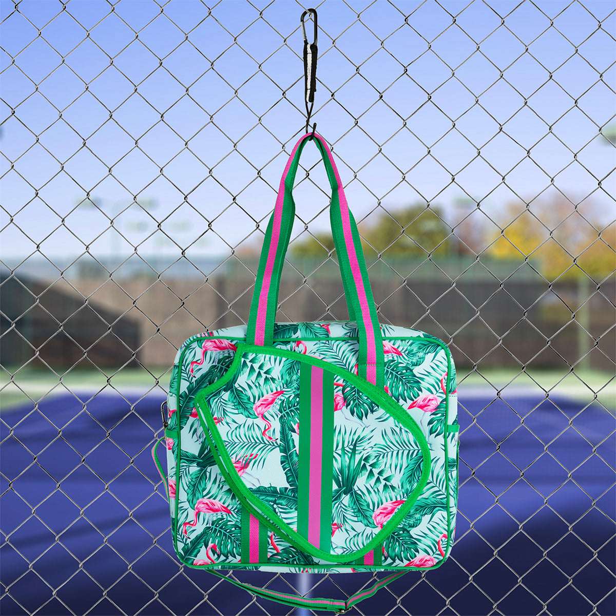 The Queen of the Court Brooklyn Pickleball Bag, featuring green, pink, and white flamingo and palm leaf designs, hangs on a fence by a blue and green tennis court with trees and clear sky in the background.