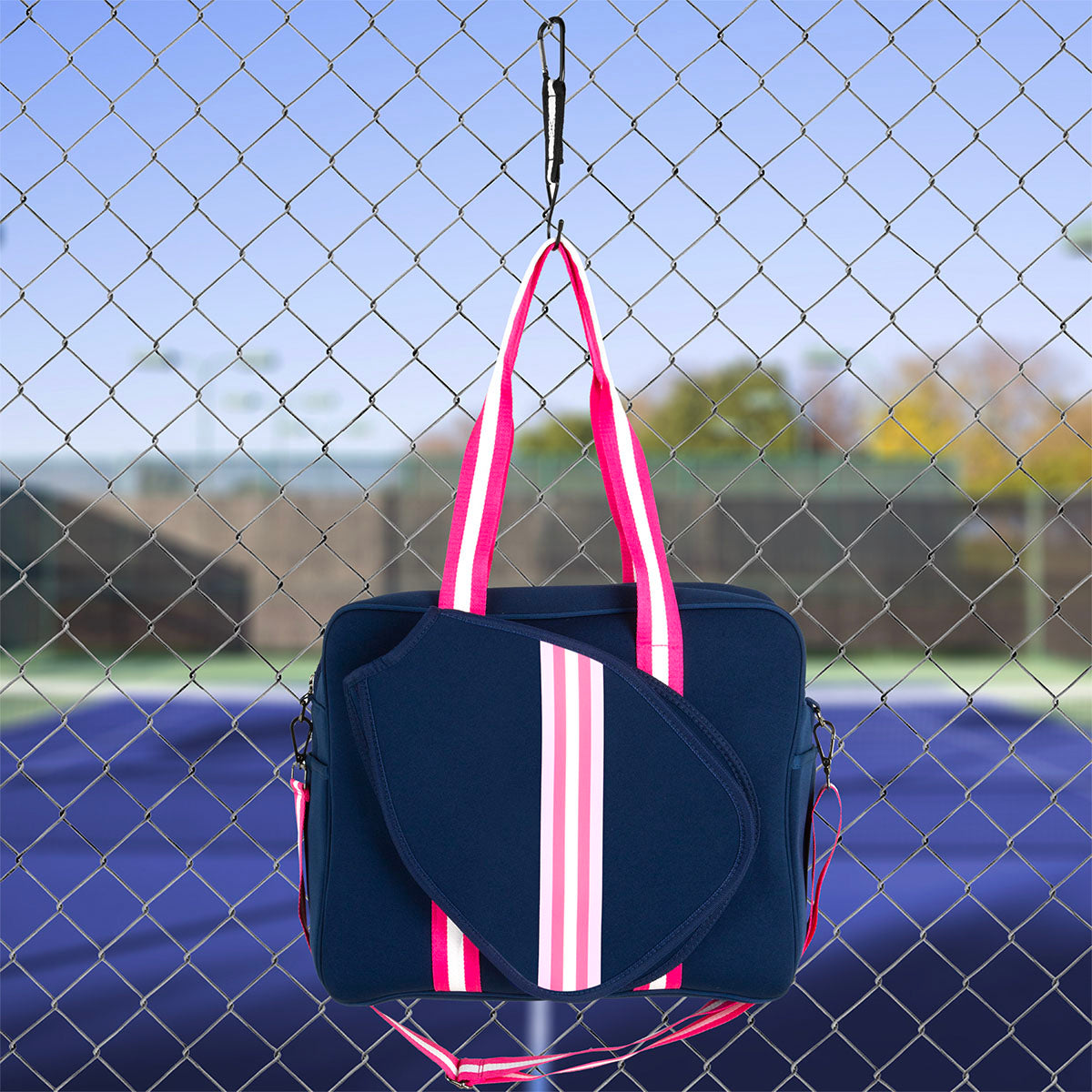 The Queen of the Court Brooklyn Pickleball Bag in navy blue, with bright pink and white striped handles and matching trim, hangs by a hook on a chain-link fence near a tennis court under a blue sky.