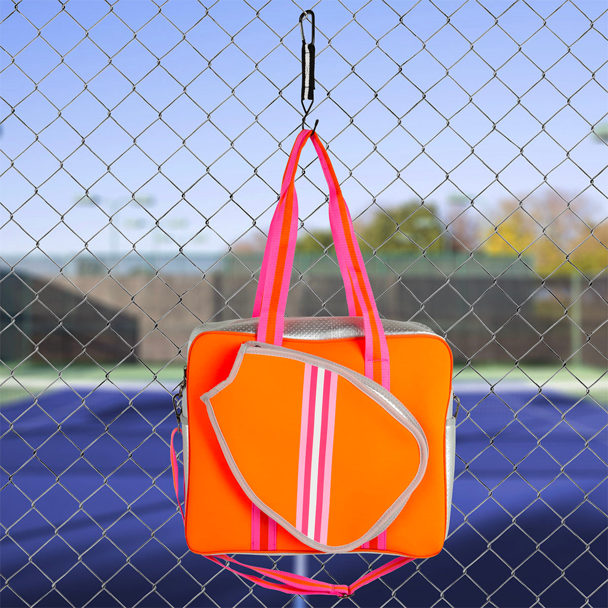 The Queen of the Court Brooklyn Pickleball Bag, in bright orange with pink and white stripes and matching straps, hangs by a black hook on a fence, its racket-shaped compartment popping against a tennis court under a clear sky.