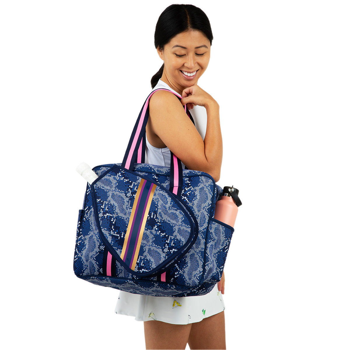 A woman with dark hair in a ponytail smiles while carrying the Queen of the Court Brooklyn Pickleball Bag—a blue and white snakeskin duffel with pink-striped handles, rainbow stripe, and two side pockets holding water bottles.