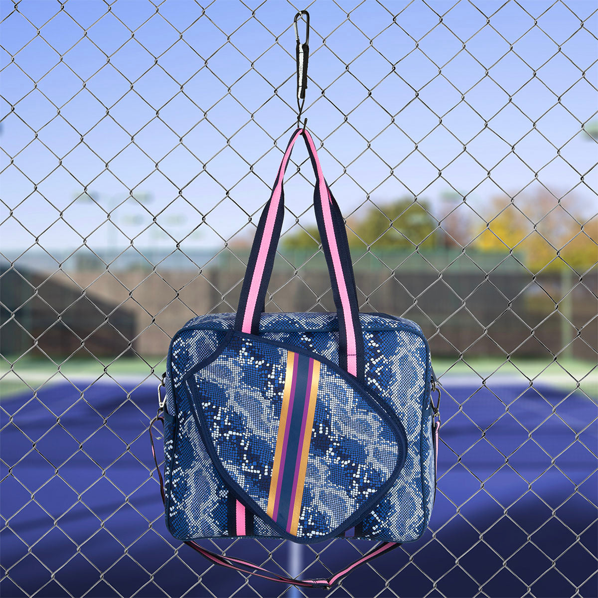 The Queen of the Court Brooklyn Pickleball Bag, featuring a blue snakeskin pattern and pink, yellow, and navy striped straps, hangs on a chain-link fence with a tennis court and blurred trees in the background.