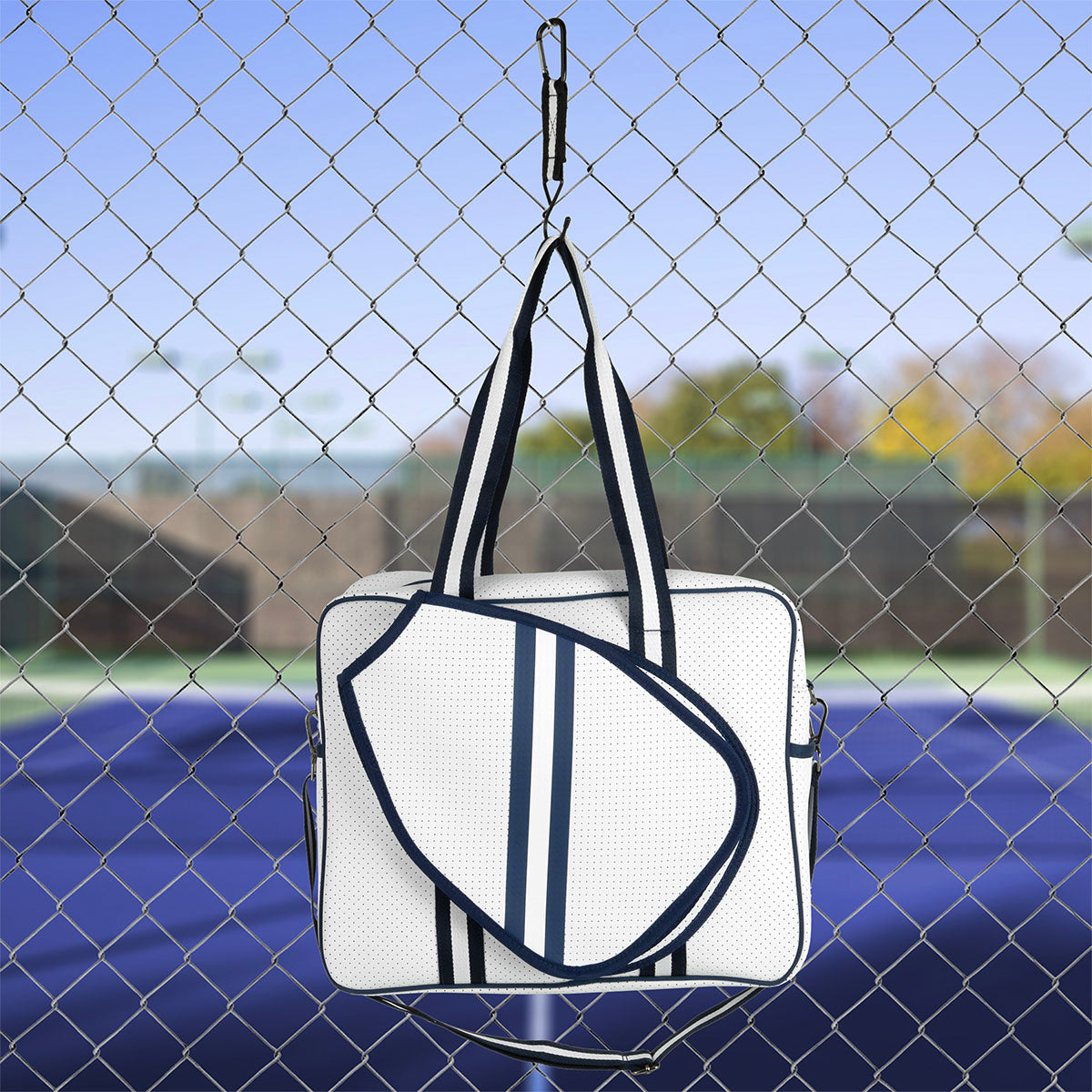 A white Queen of the Court Brooklyn Pickleball Bag with blue stripes, long handles, and an attached racquet cover hangs on a fence by a tennis court. The background features a net, green court, and blurred trees under a blue sky.