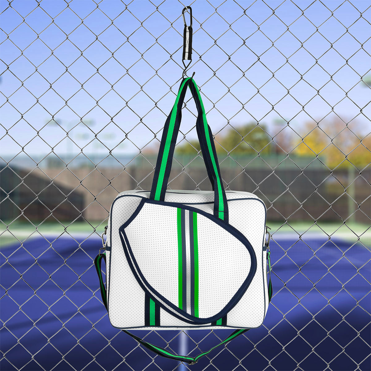 A white, rectangular Queen of the Court Brooklyn Pickleball Bag with green and navy stripes, two handles, and a matching shoulder strap hangs on a chain-link fence; tennis courts and blurred trees are visible in the background.
