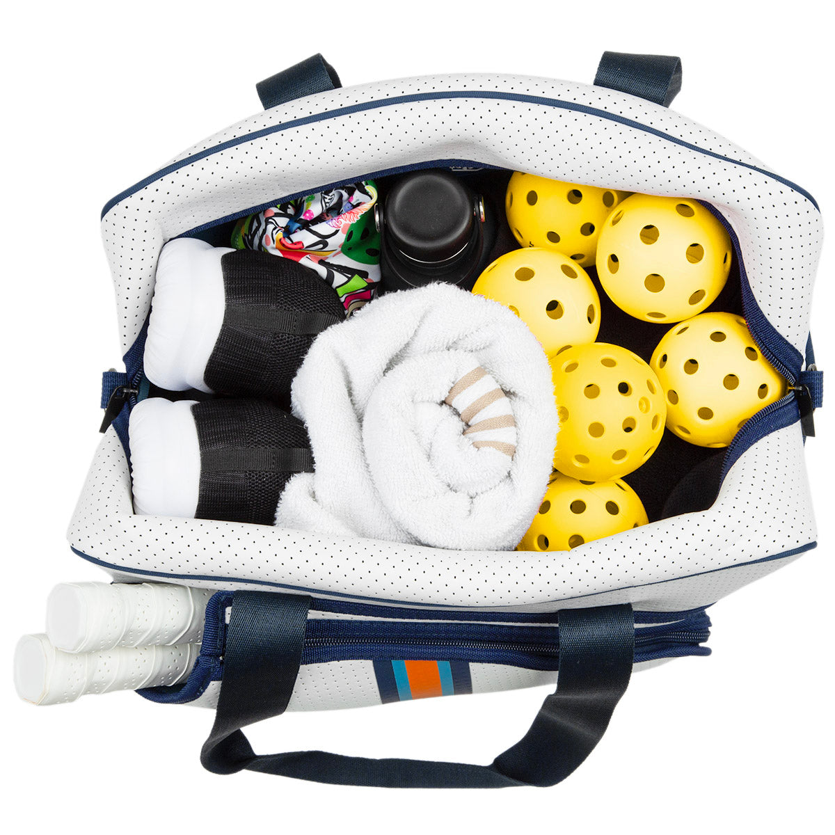 A top-down view of the Queen of the Court Brooklyn Pickleball Bag displays yellow pickleballs, white towels, water bottles, black-and-white sports socks, and three white-handled paddles. The bag features navy straps and a white exterior with blue and orange stripes.