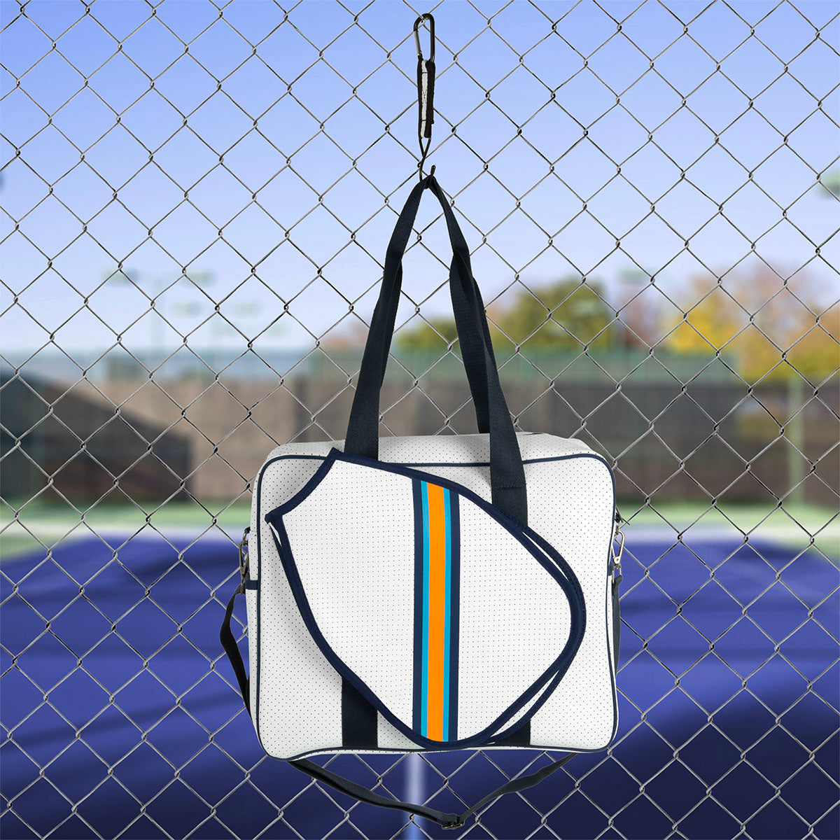 The Queen of the Court Brooklyn Pickleball Bag, featuring black straps and blue, orange, and yellow stripes, hangs on a chain-link fence with a tennis court and trees blurred in the background under a clear sky.