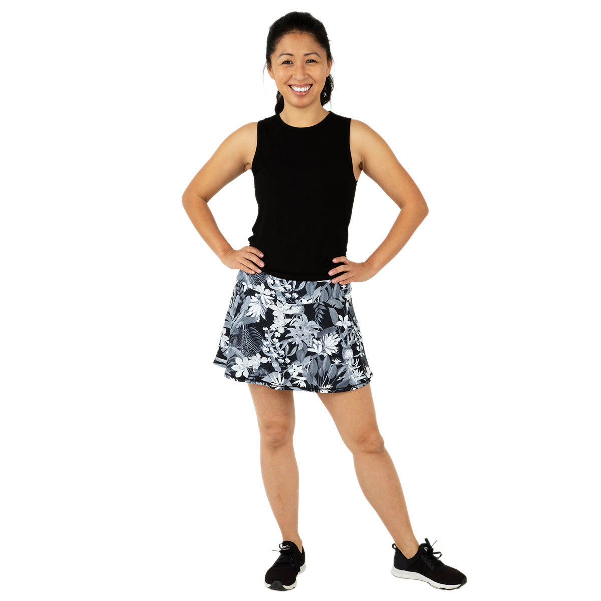 A smiling woman stands with hands on hips, wearing the Queen of the Court Tropical Swing Skirt and a sleeveless black top. Her outfit, paired with black athletic shoes, highlights stylish pickleball activewear against a plain white background.
