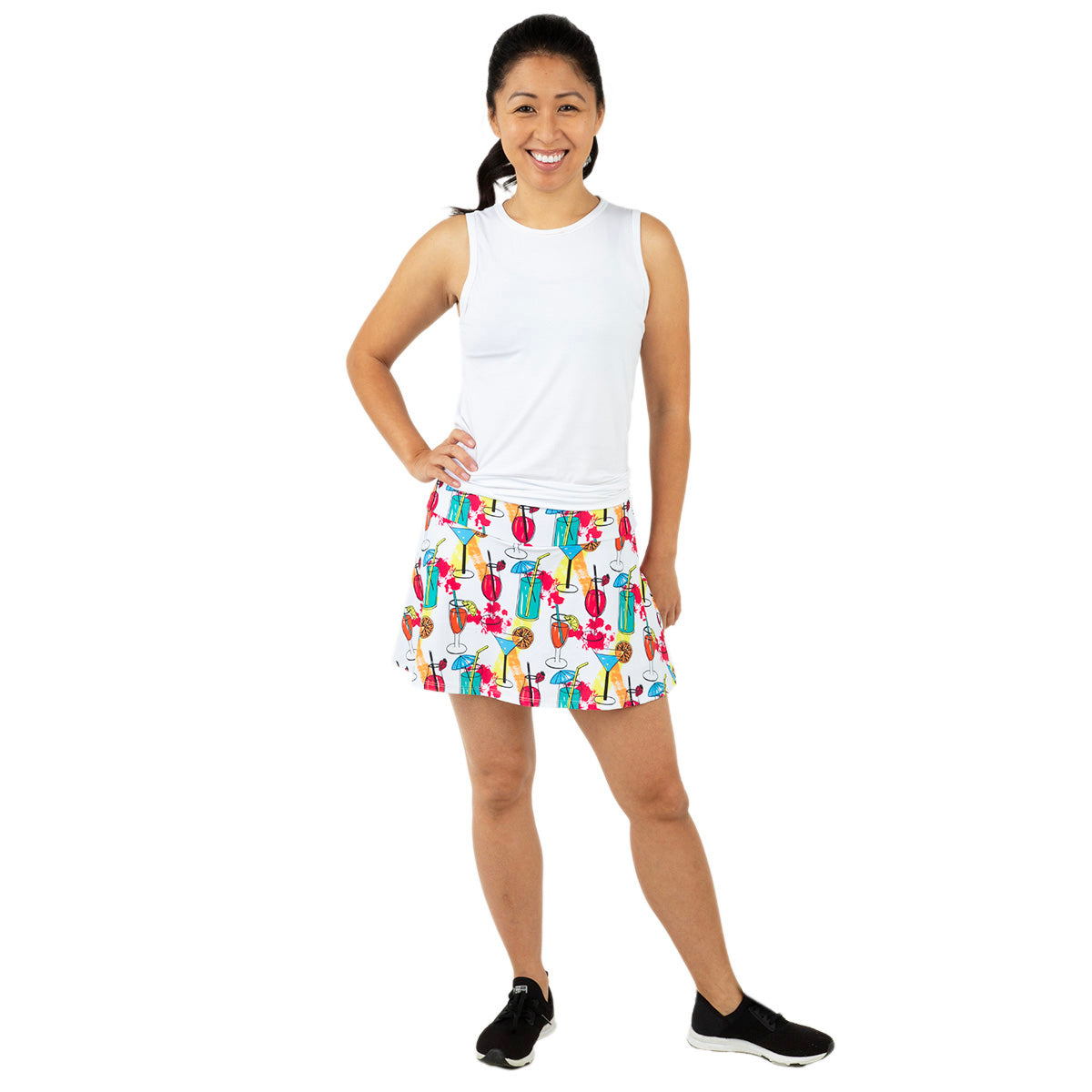 A woman smiles in the Queen of the Court Cocktails on the Court Swing Skirt, a colorful tennis skirt with golf bag and club patterns, paired with a sleeveless white top and black sneakers. She stands on a white background, hand on hip.