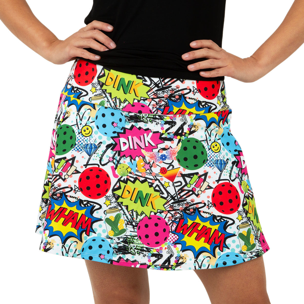 A person stands with hands on hips, wearing the Queen of the Court Dink Dink Wham Swing Skirt—an active skirt with comic-style DINK, WHAM, paddles, pickleballs, and smiley faces—paired perfectly with a black top.